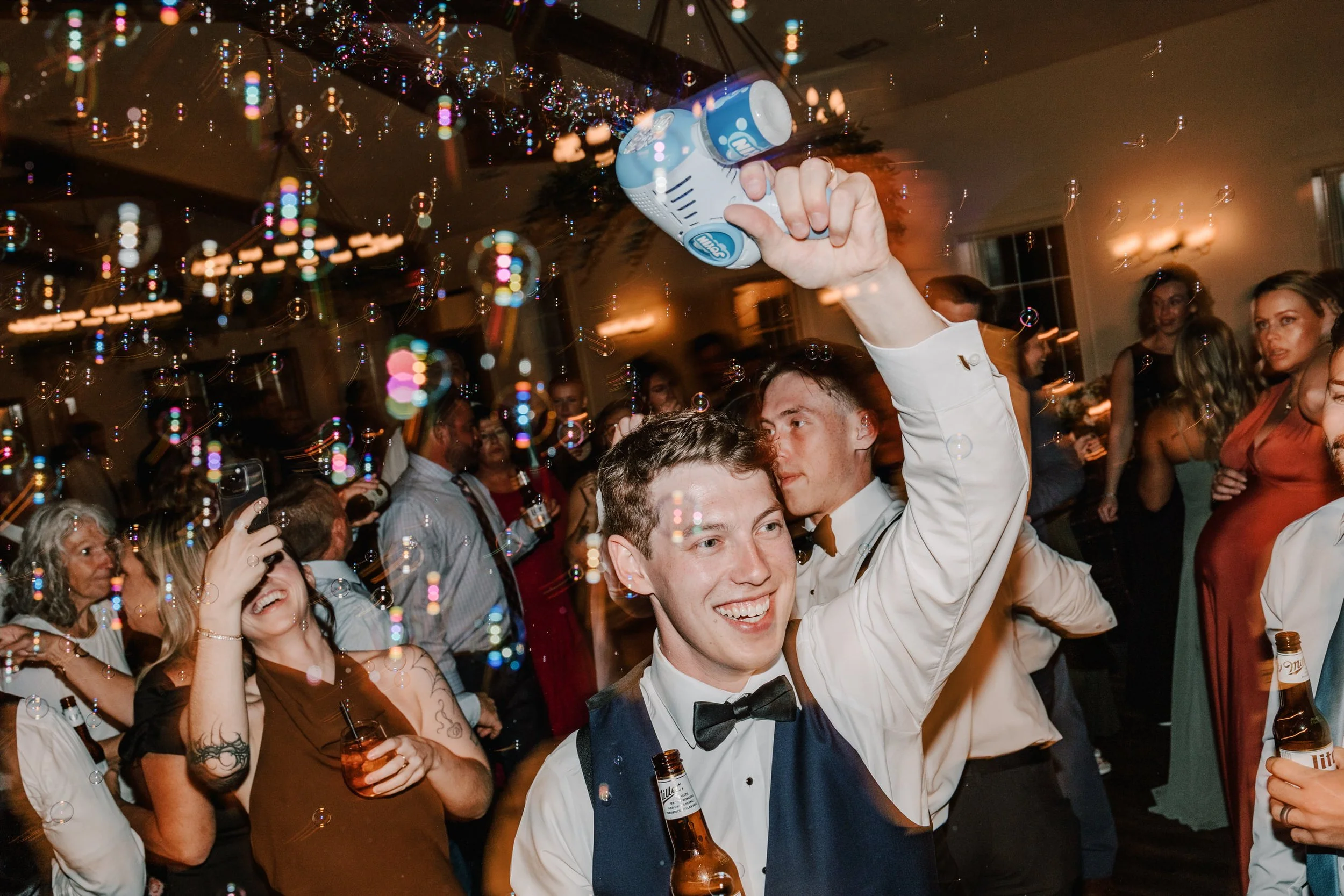 People dancing and celebrating at a party, with a young man holding a spray can and smiling, surrounded by others, some holding drinks, with balloons and colorful confetti in the air.