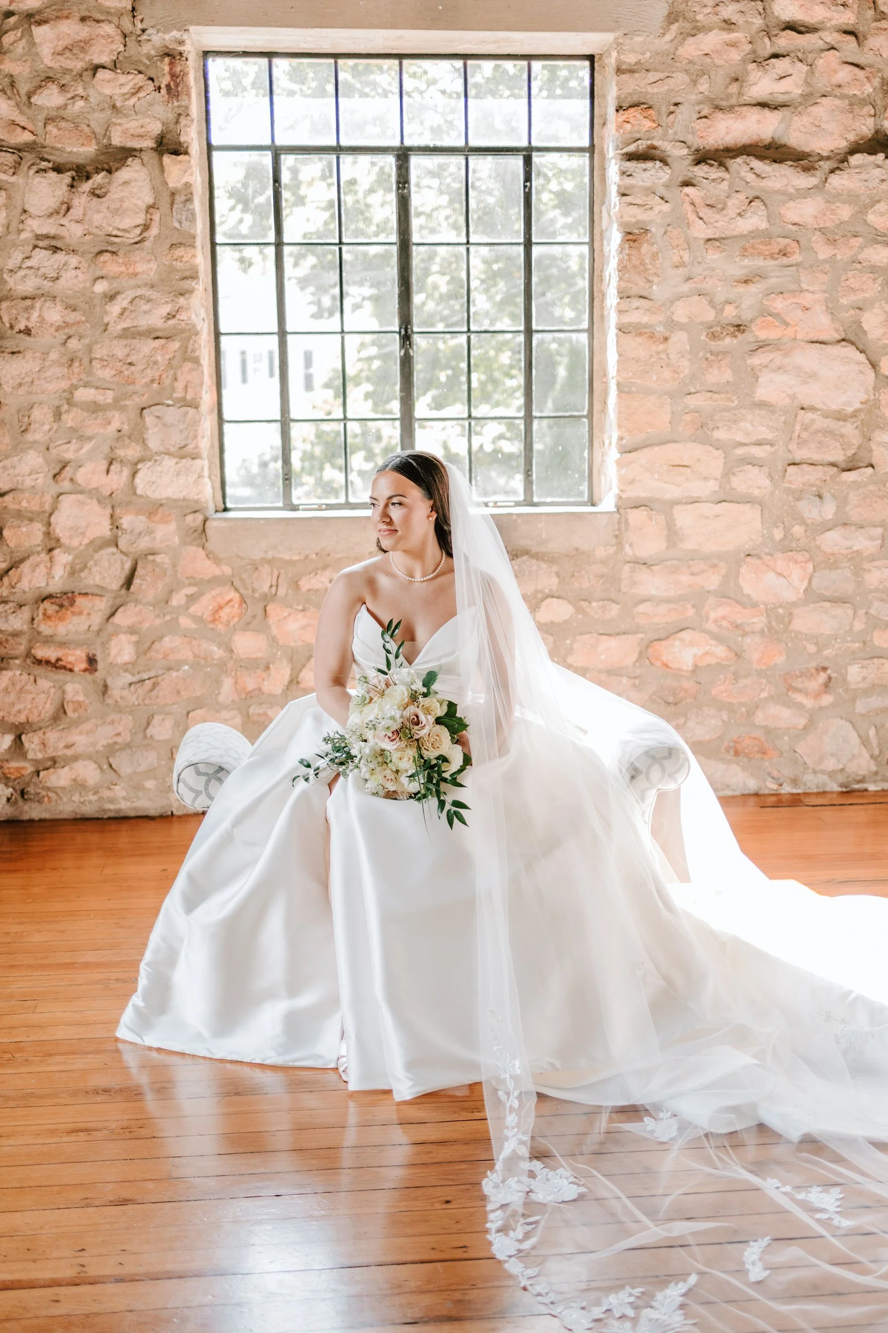 Bride sitting on a bench in a rustic brick-walled room with a large window, wearing a white wedding dress and holding a bouquet of white and blush roses, with a long veil flowing on the floor.