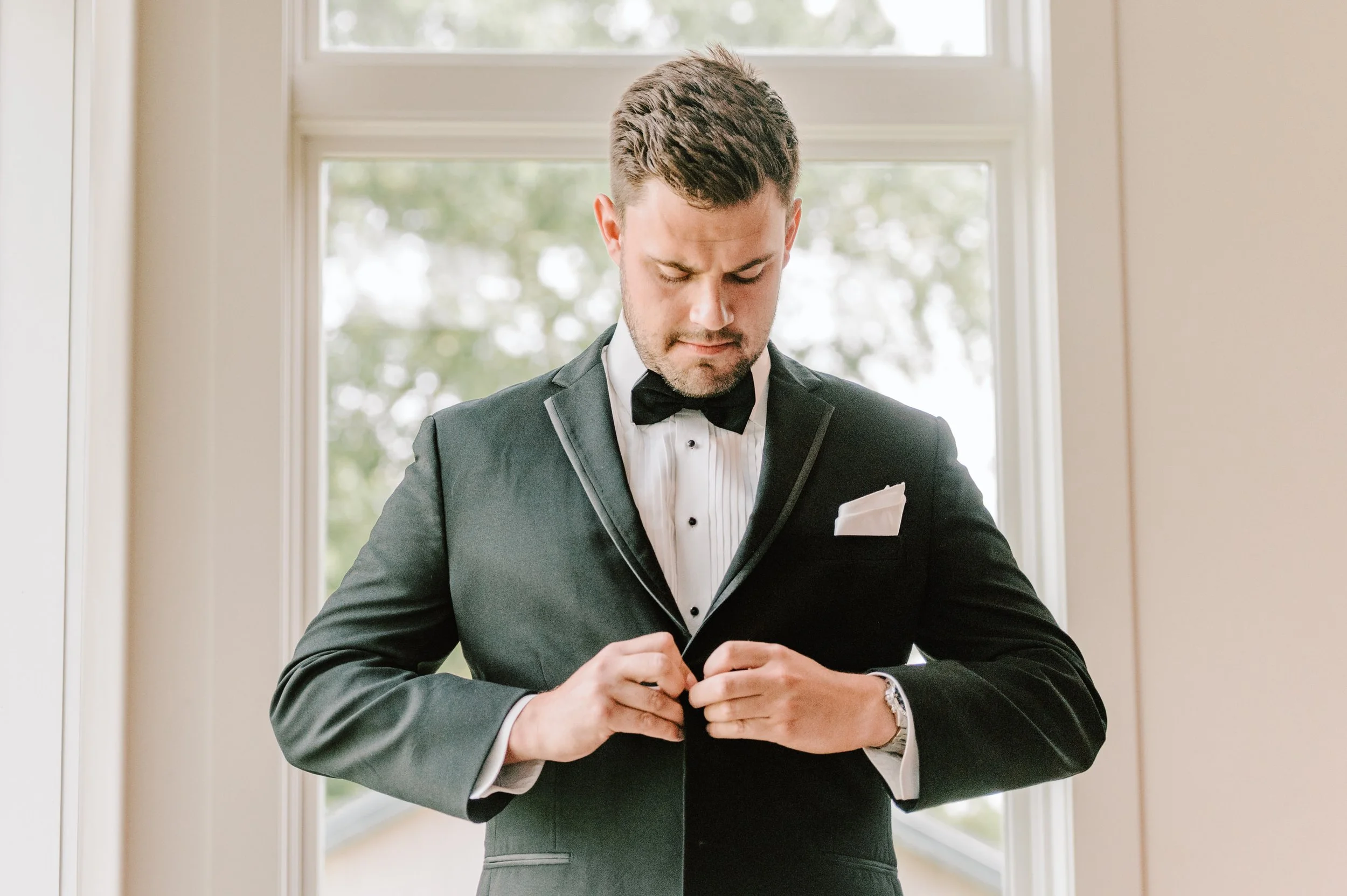 A man in a tuxedo with a white dress shirt and black bow tie is buttoning his jacket, standing in front of a window with trees outside.