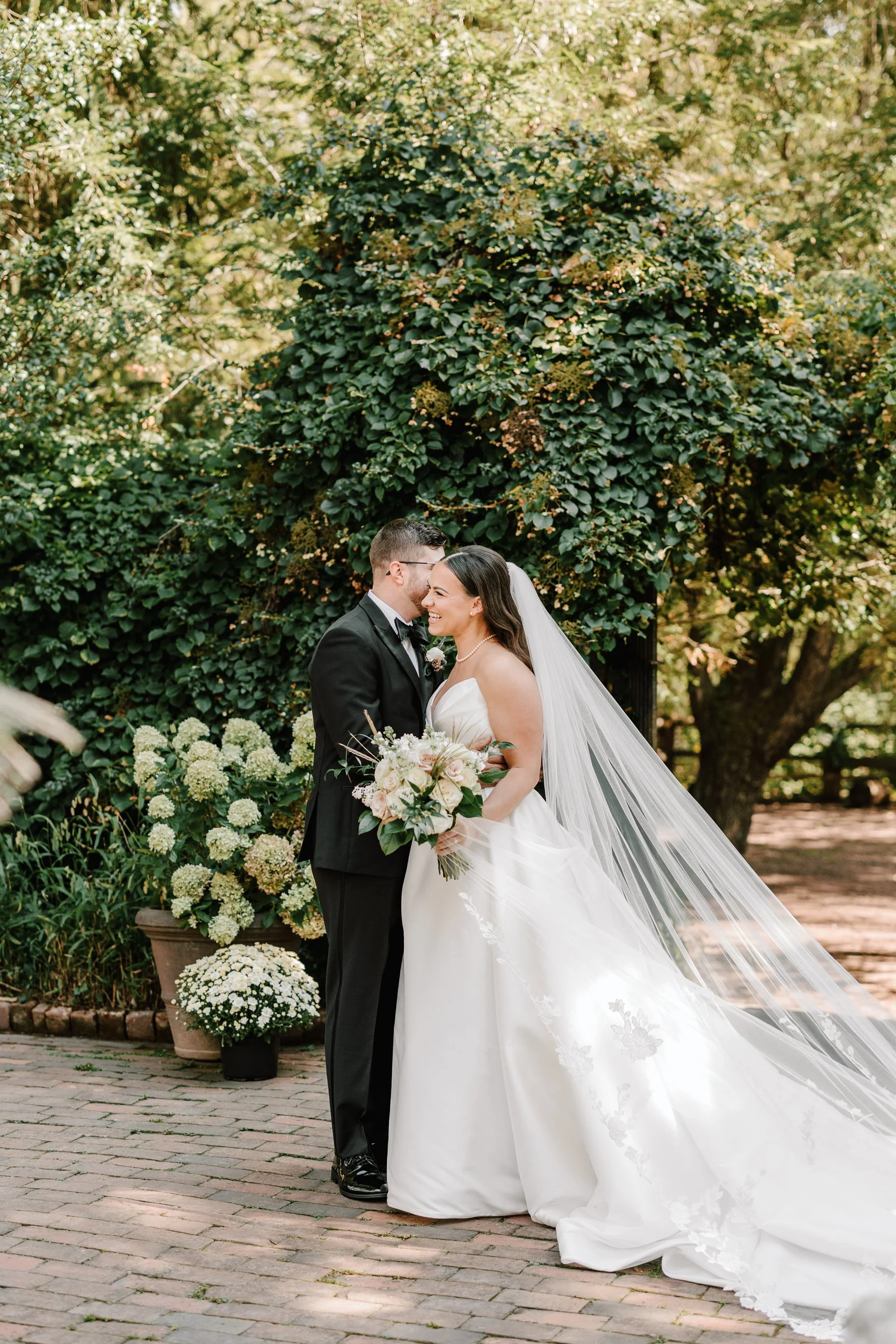 A bride and groom are standing close together outdoors, smiling, with the bride holding a bouquet of flowers. The bride is wearing a white wedding dress with a long veil, and the groom is dressed in a black tuxedo. The background features greenery an