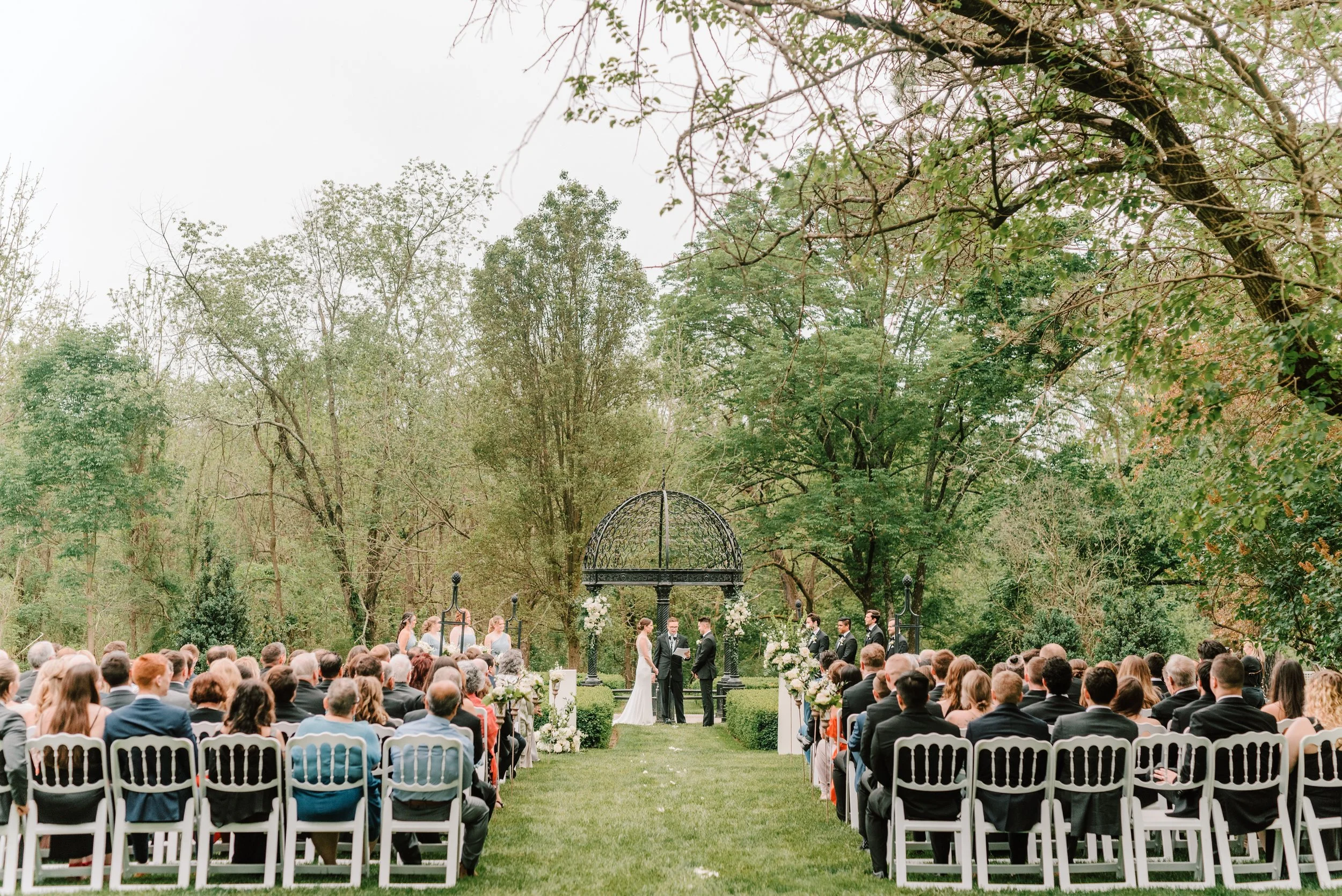 Outdoor wedding ceremony with a bride and groom exchanging vows under a black metal arch, surrounded by seated guests on white chairs, in a lush green park with trees and overcast sky.