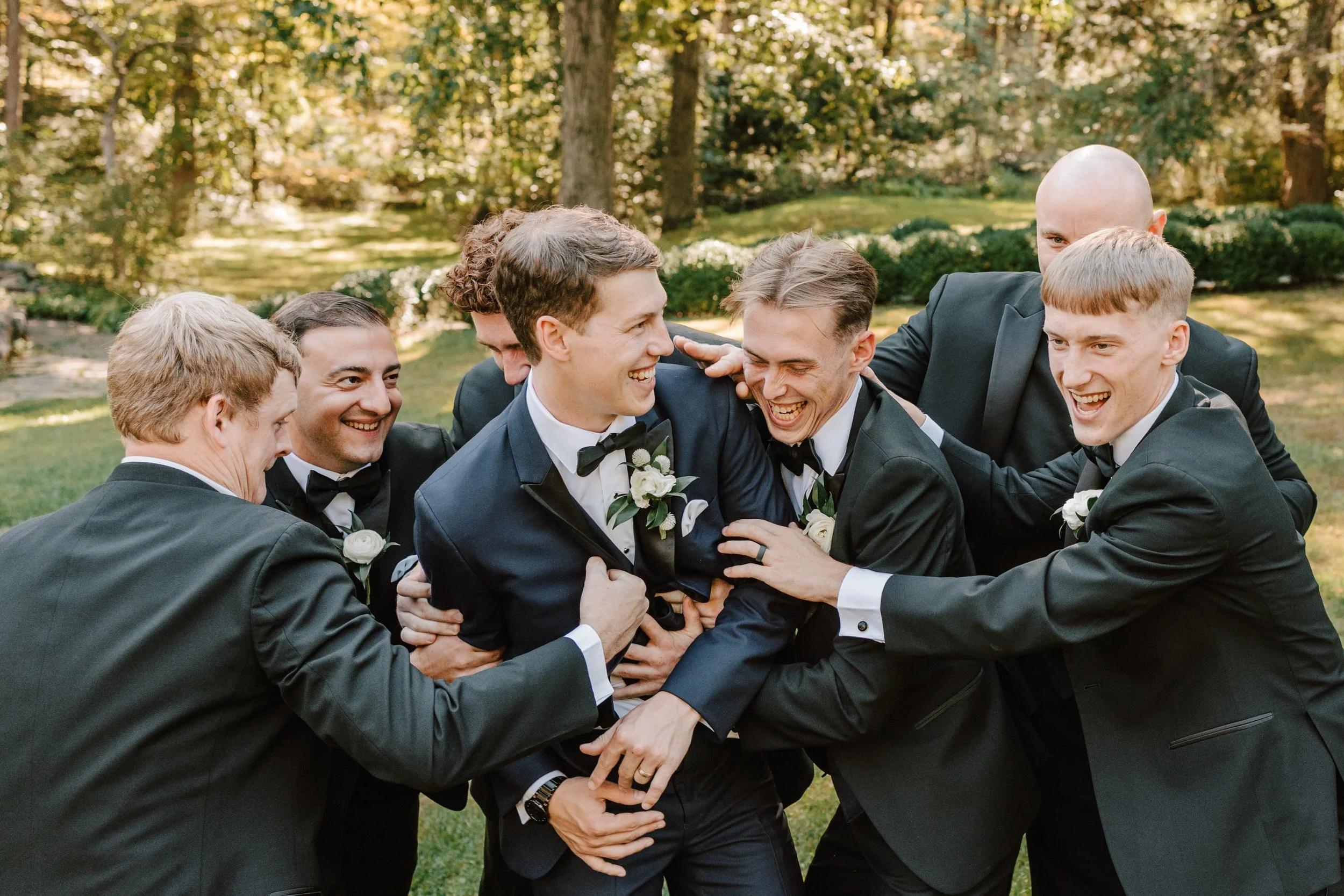 Group of men in tuxedos laughing and smiling outdoors in a garden setting.
