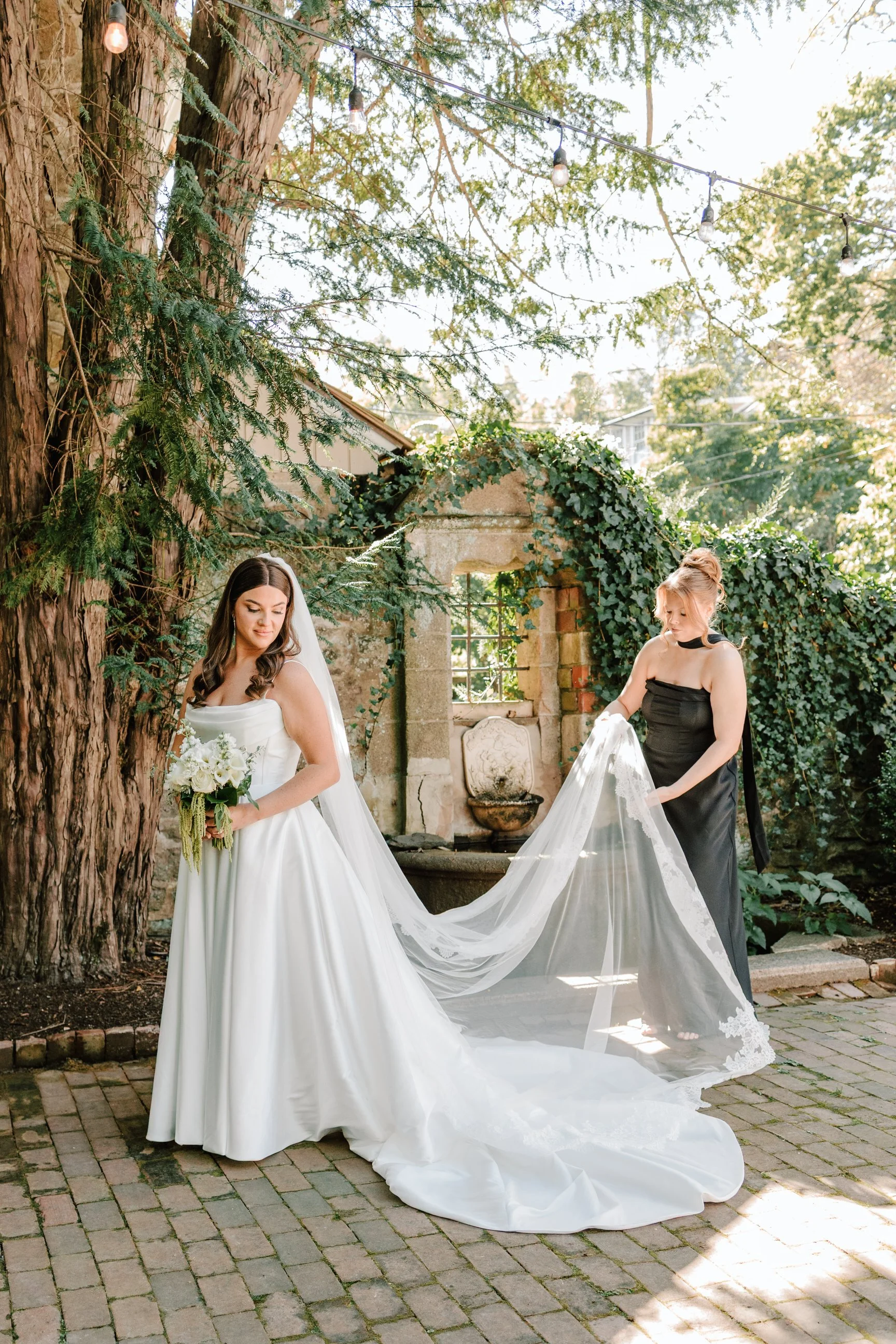 Bride in wedding dress holding bouquet, standing outdoors with a woman in black dress adjusting her train, lush greenery and stone outdoor fountain in background.