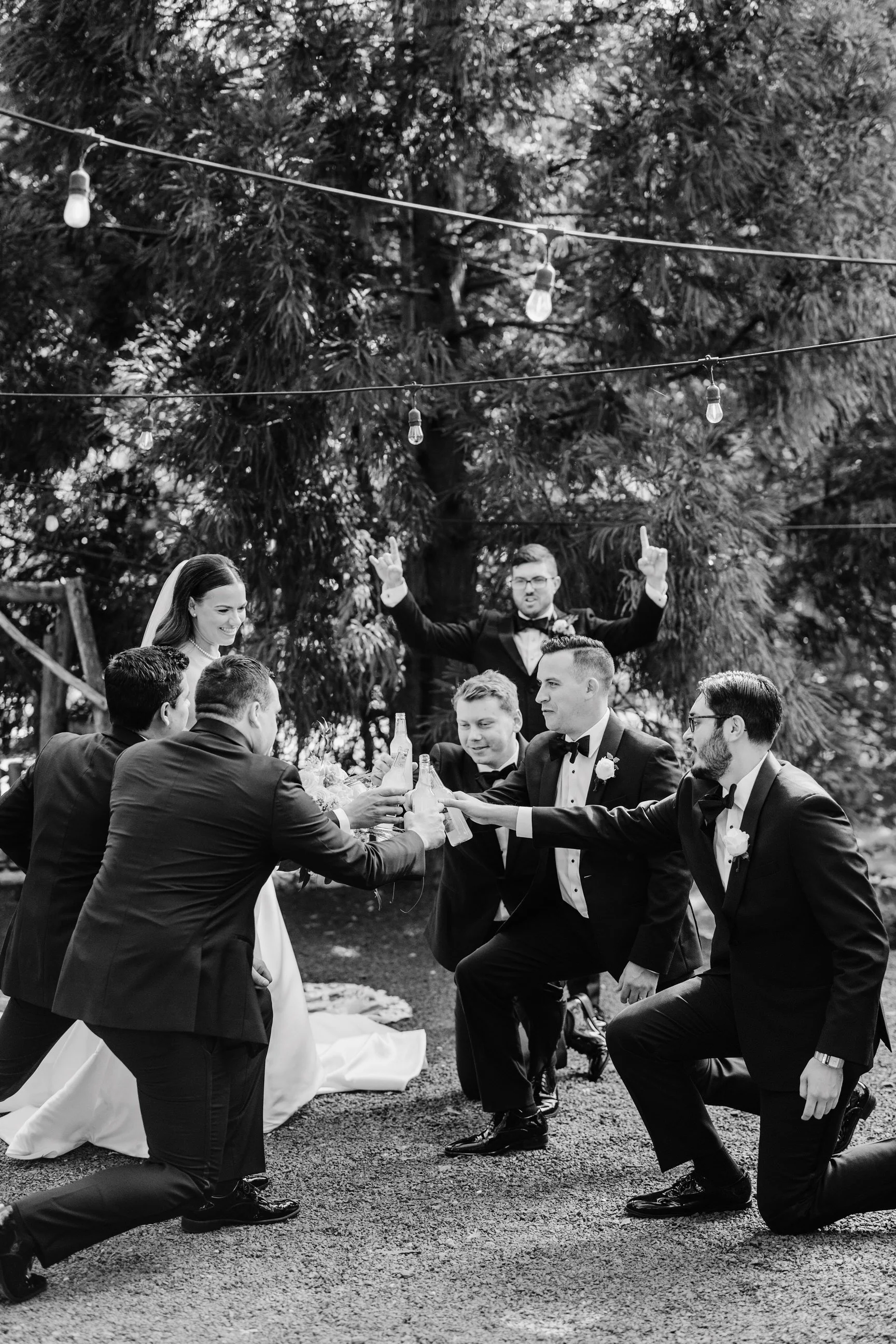 Black and white photo of a wedding toast outdoors with the bride, groom, and groomsmen raising drinks in a celebratory toast, under string lights, with a large tree in the background.