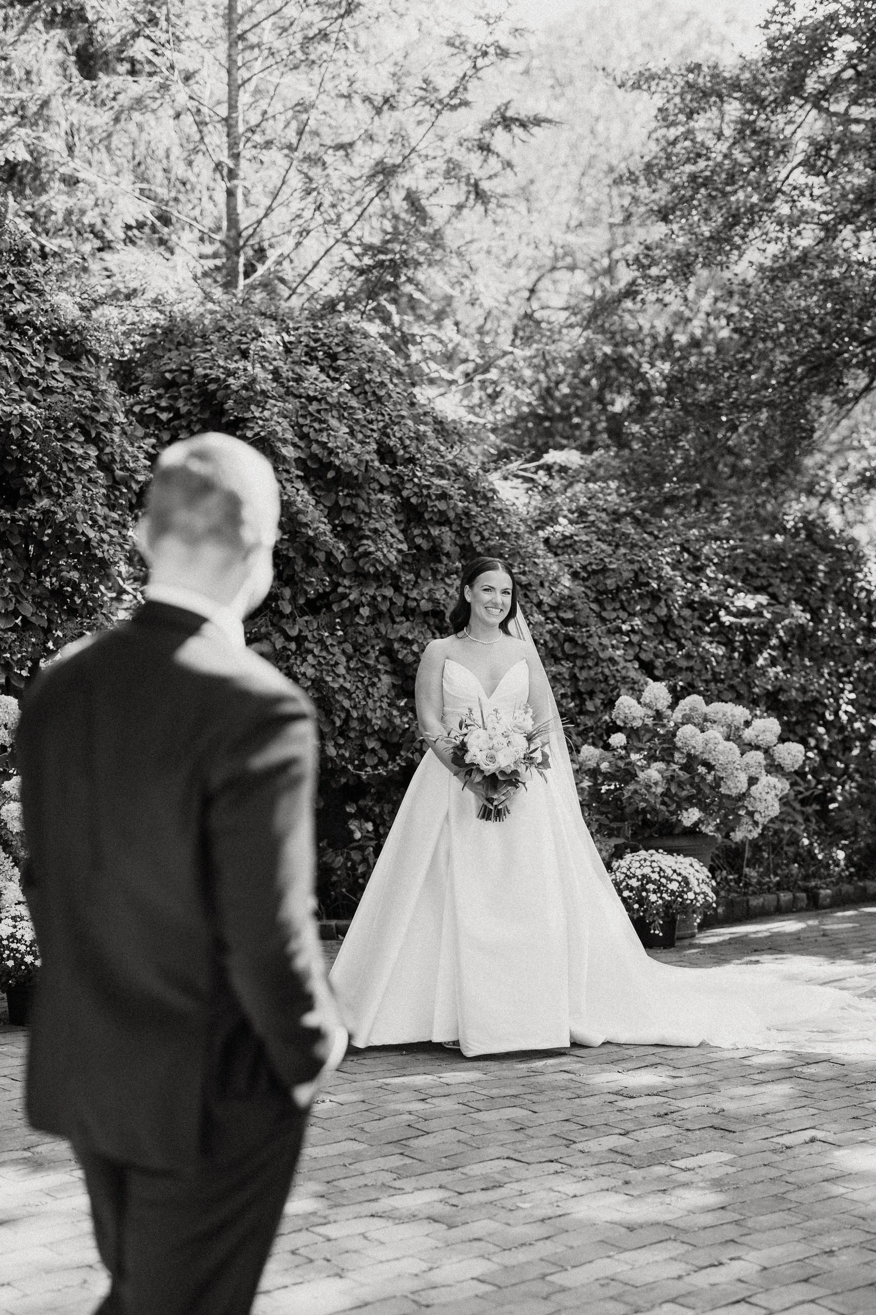 A bride in a white wedding gown holding a bouquet of flowers, smiling as she looks at a person in a suit, in an outdoor garden setting.