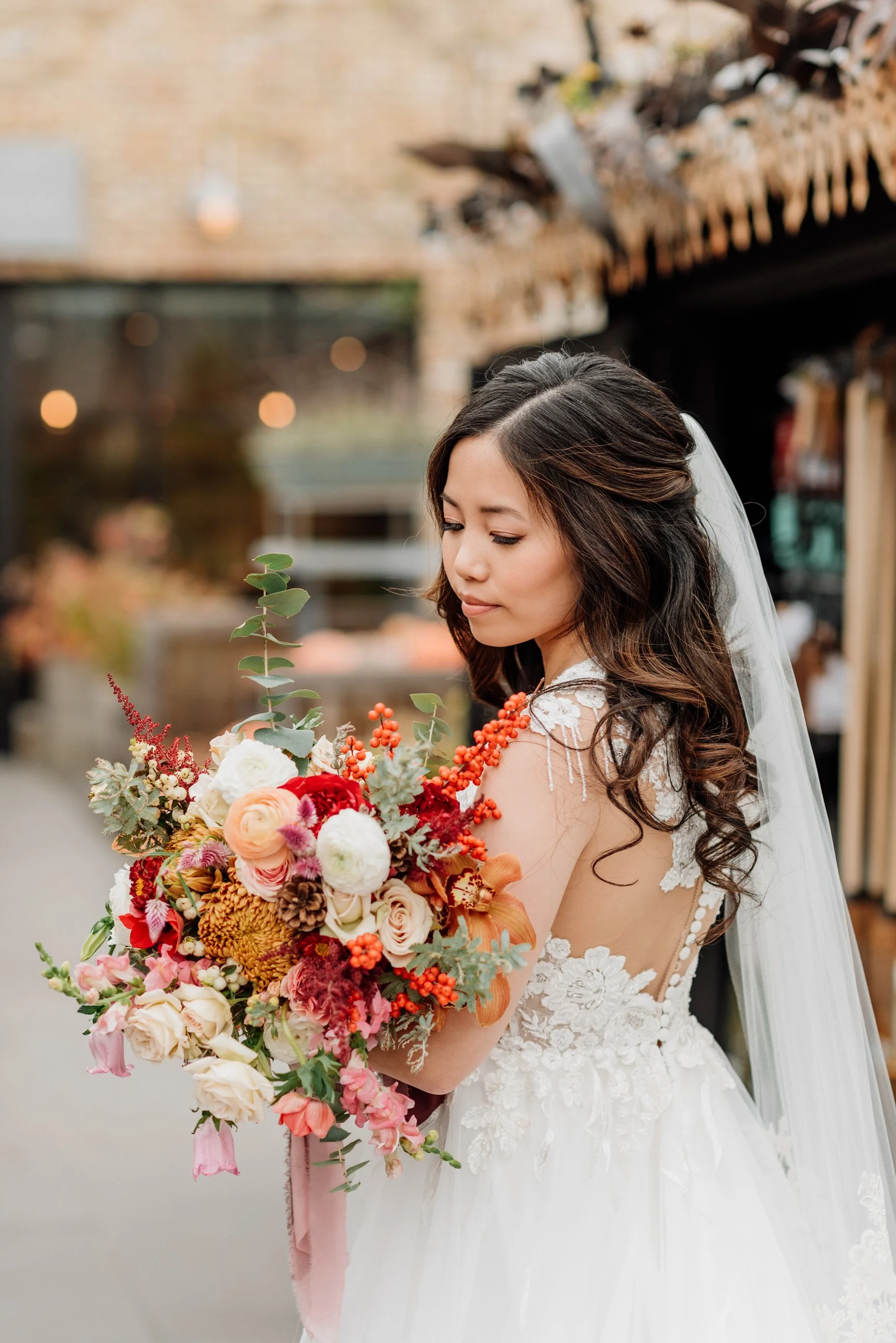 Bride in a white lace wedding dress holding a colorful bouquet of flowers, standing outdoors with traditional decorative buildings in the background.