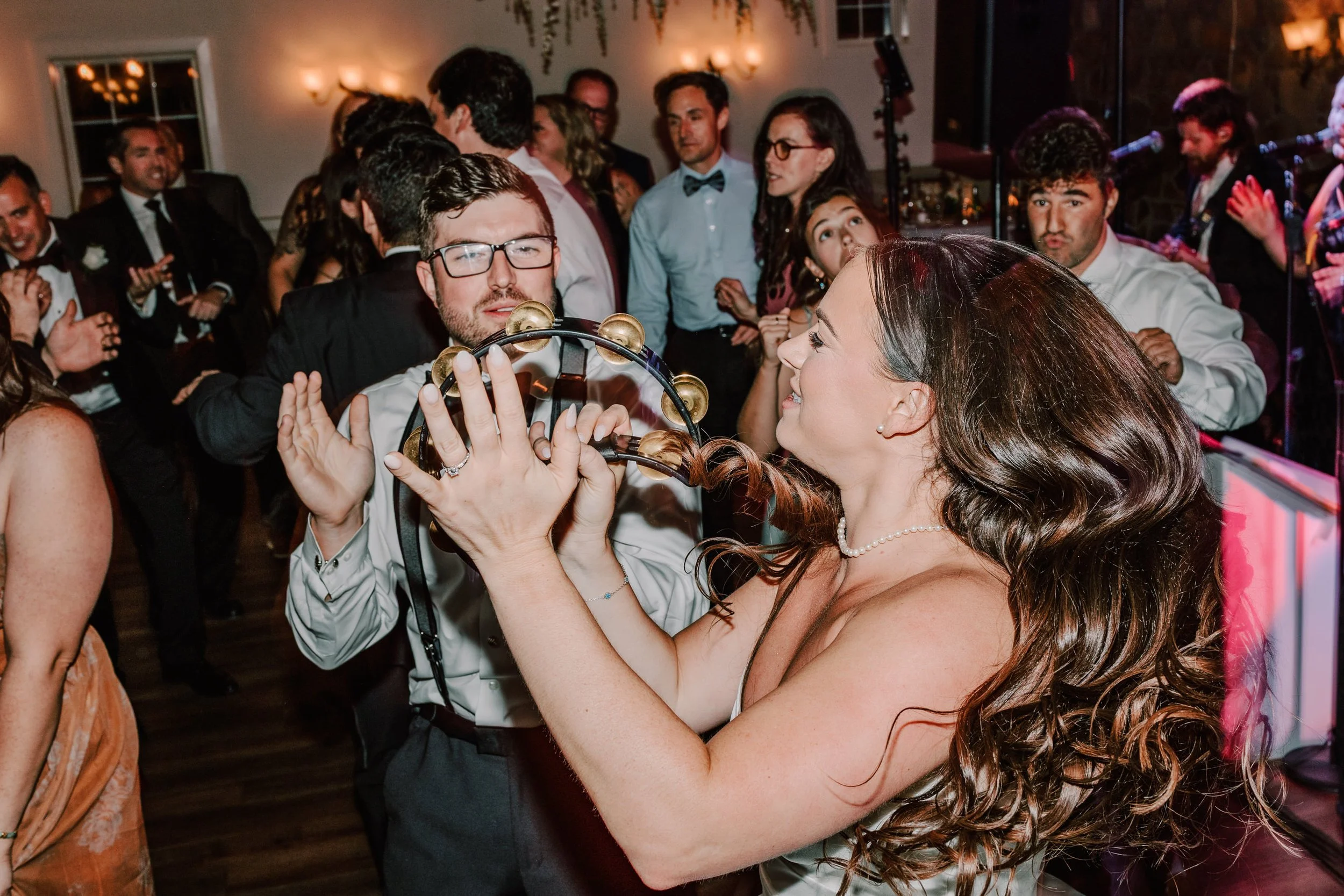 A woman with long dark hair, wearing a pearl necklace and white dress, is playing a tambourine while smiling at a man with glasses, dressed in a tuxedo. They are at a lively party or wedding reception with many people dancing and enjoying themselves 