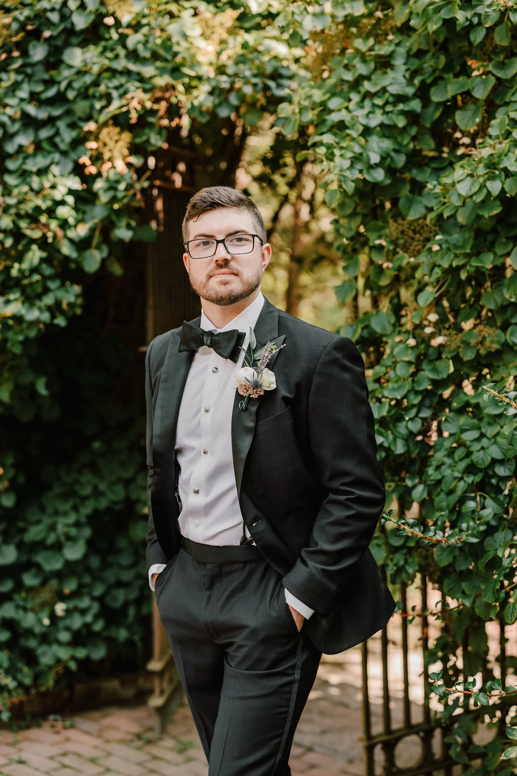 A man dressed in formal tuxedo with a boutonniere, black bow tie, and glasses standing outdoors in front of greenery.