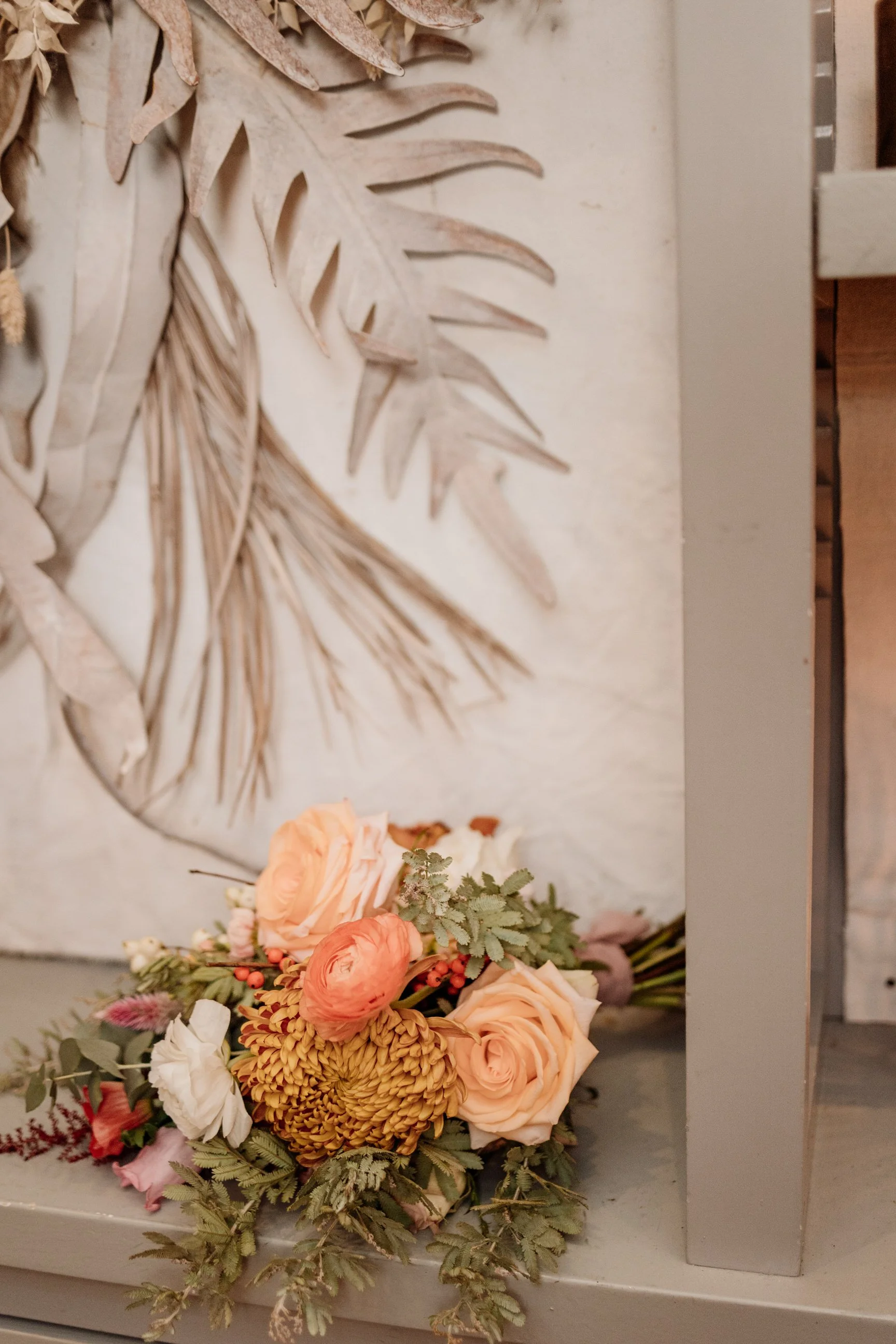 A bouquet of peach, pink, and white flowers with green foliage rests on a gray surface, with a dried monstera leaf wall art in the background.