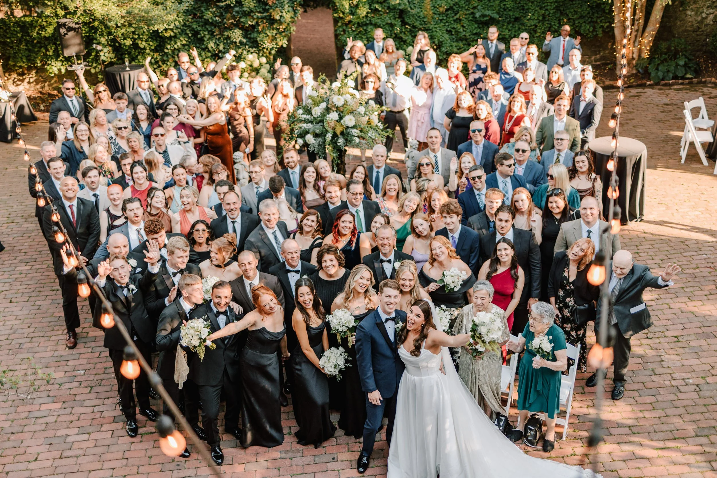 Wedding celebration with a large group of guests, including bride and groom, standing on a brick patio outdoors, surrounded by trees and string lights.