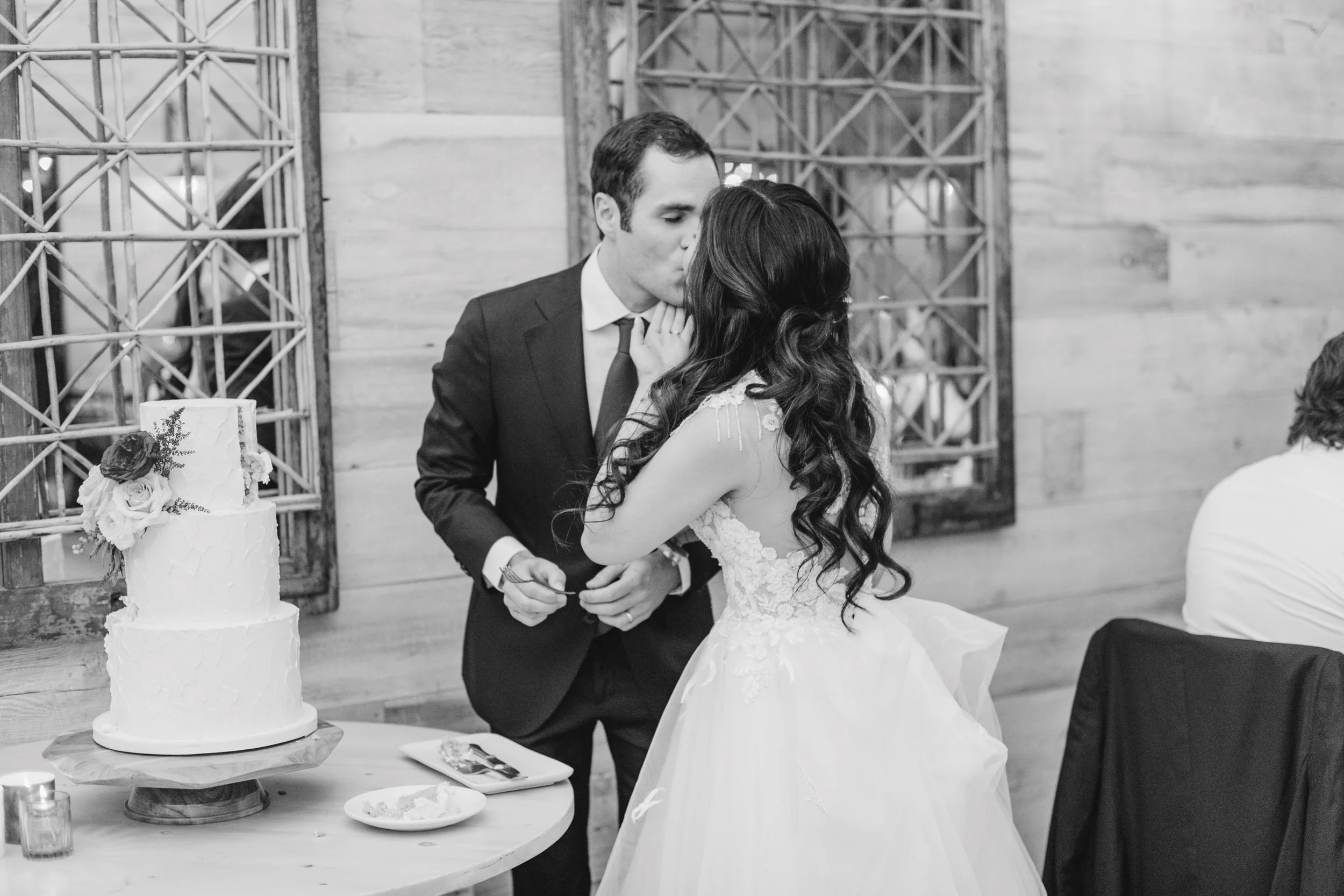 A black-and-white photo of a bride and groom sharing a kiss at a wedding reception. The bride has long, wavy hair and is wearing a lace wedding gown, and the groom is in a suit and tie. There is a wedding cake with flowers on a table nearby.