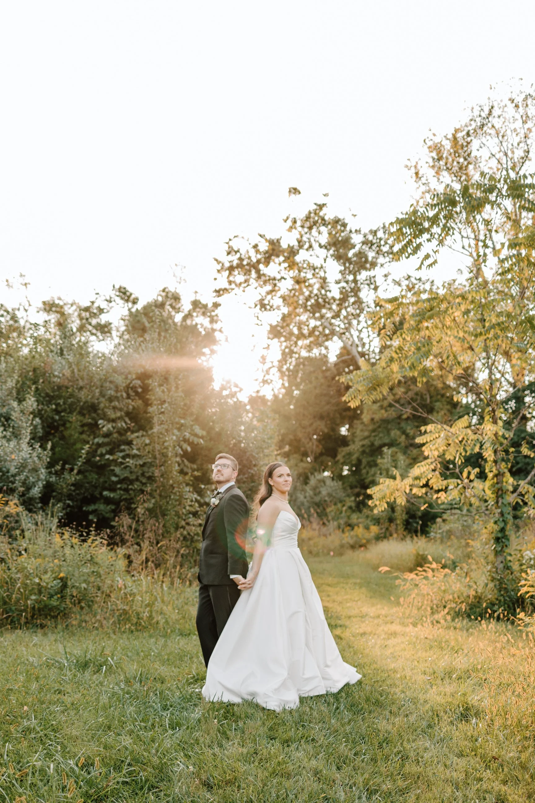 A bride and groom holding hands outside in a grassy area during sunset, surrounded by trees.