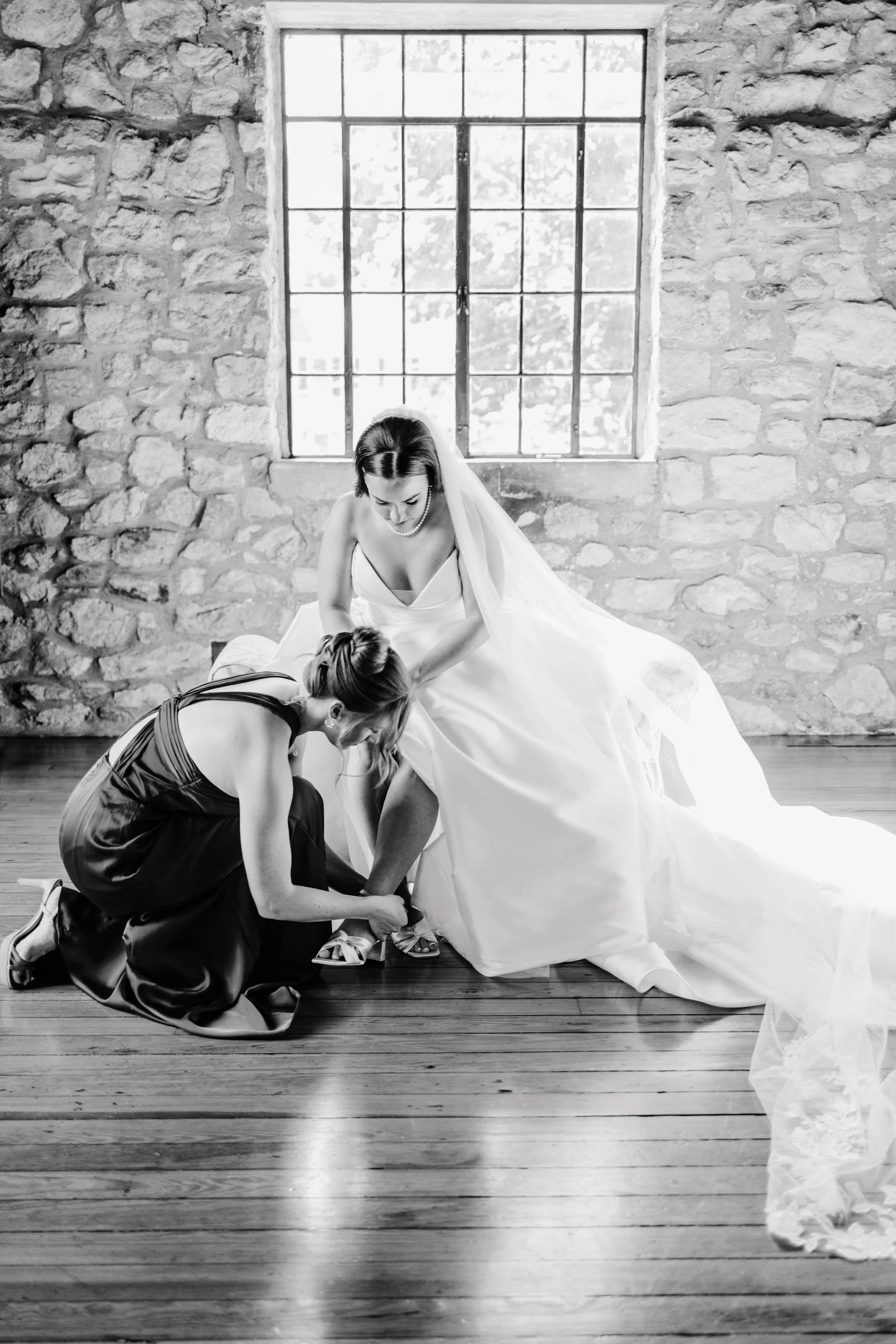 A bride in a wedding dress is assisted with shoes by another woman in a black gown, inside a room with a large window and stone wall.