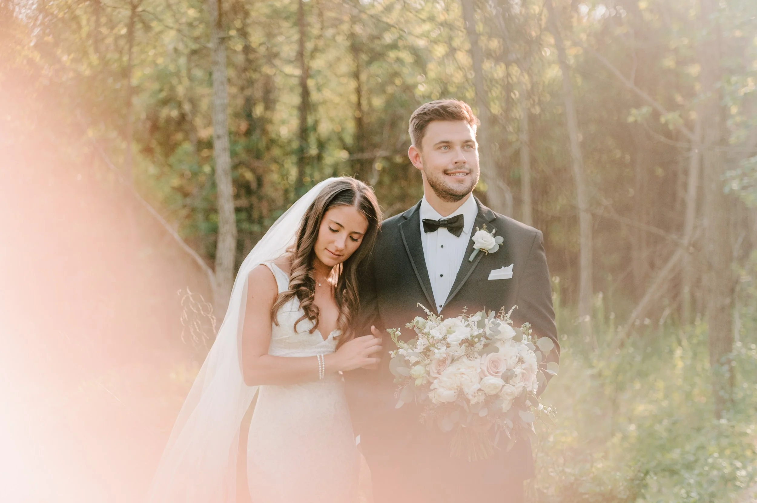 Bride and groom standing outdoors in a wooded area during a wedding ceremony, with the groom holding a bouquet of flowers.