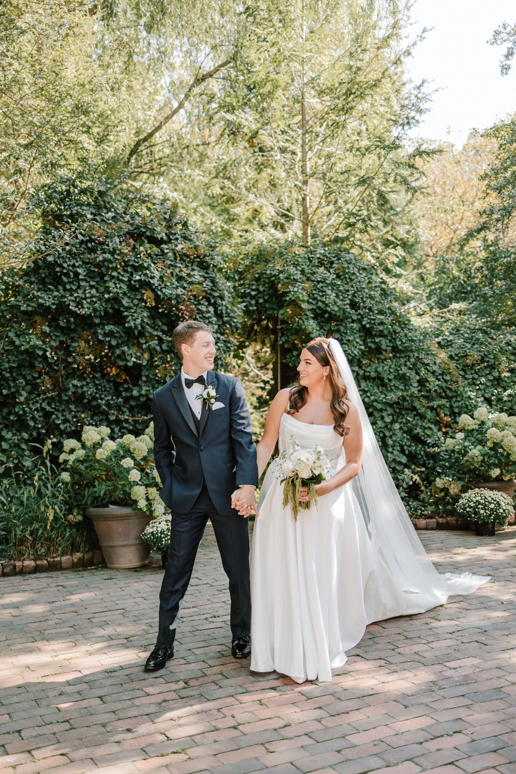 A bride and groom holding hands and smiling at each other outdoors on a brick pathway surrounded by lush green trees and plants.
