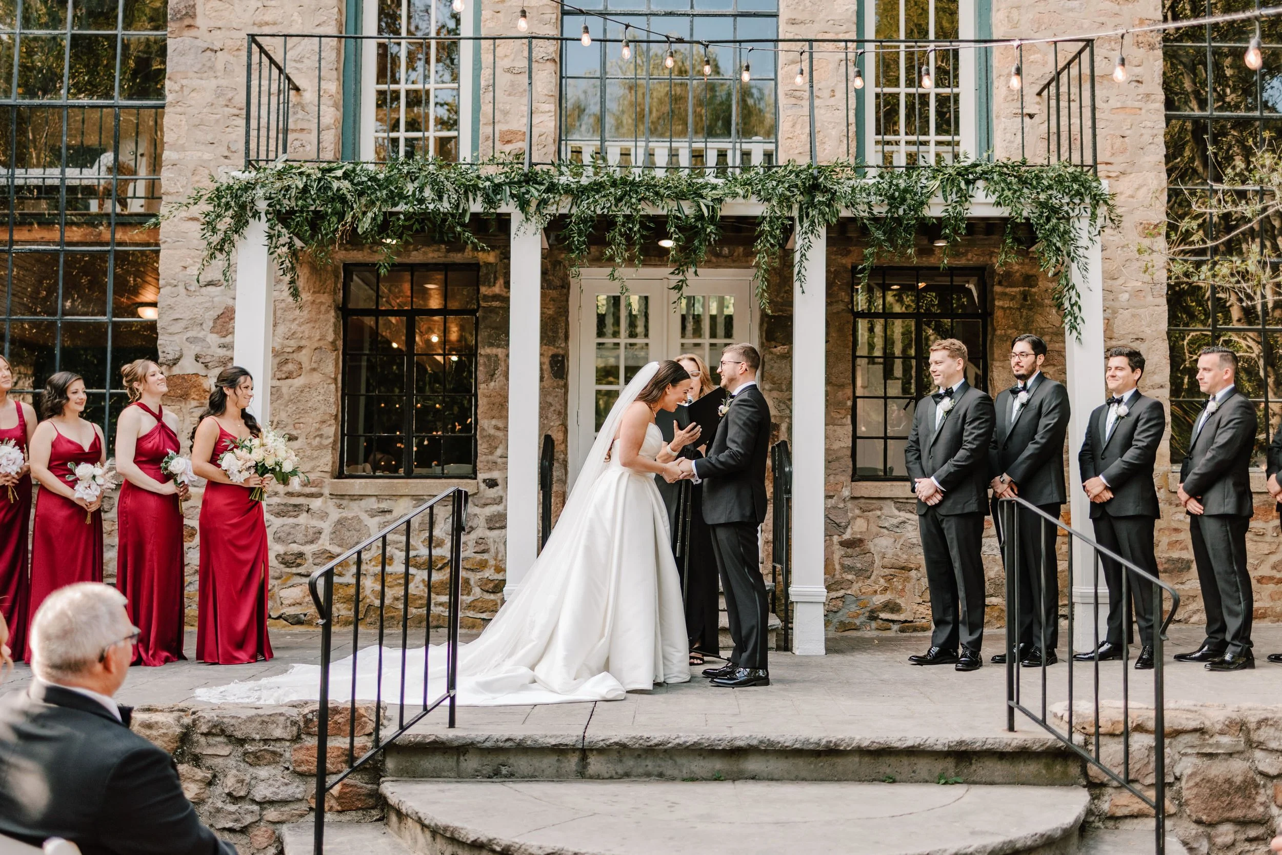 Wedding ceremony with bride and groom holding hands, bride in white gown and veil, groom in black tuxedo, officiant reading, bridesmaids in matching red dresses holding bouquets, groomsmen in black tuxedos, stone building backdrop with fairy lights a