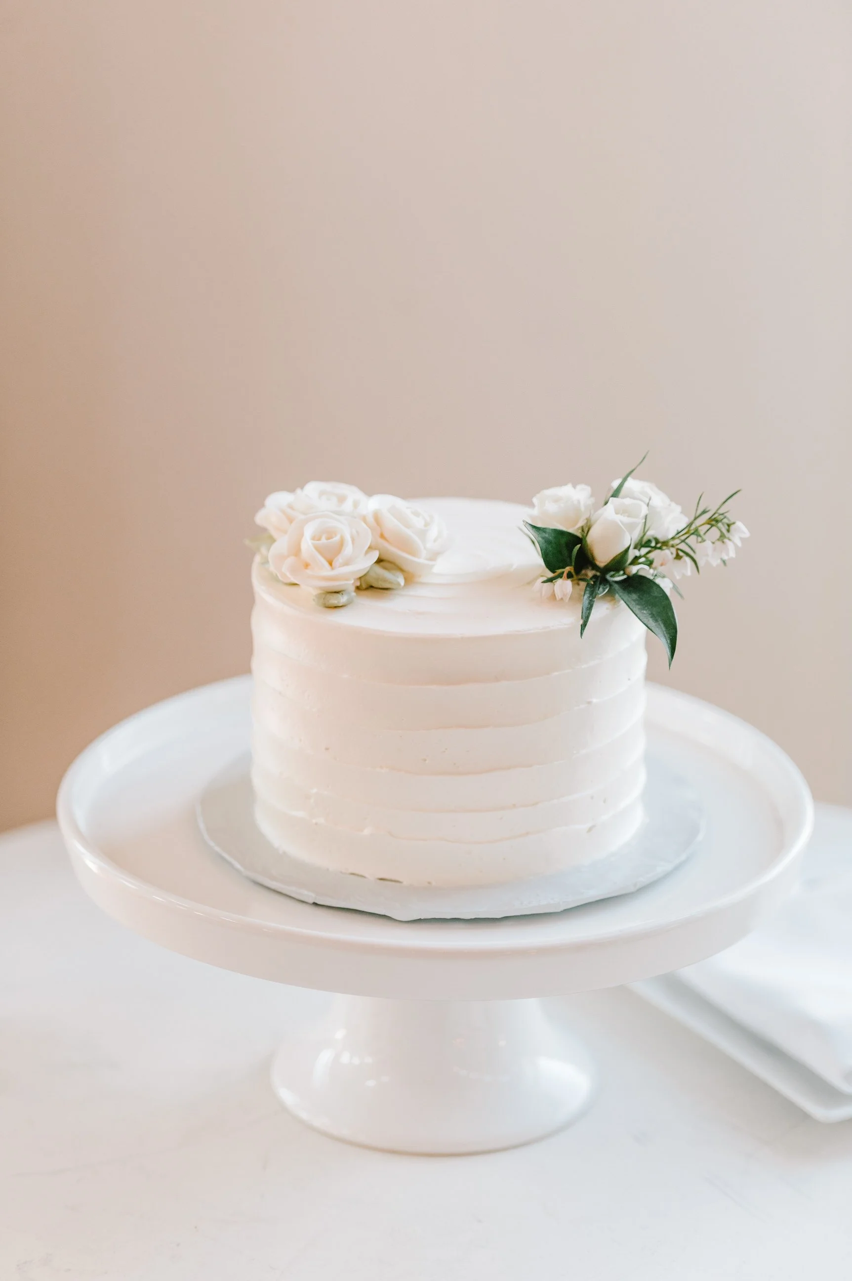 A white wedding cake decorated with white roses and green leaves on a white cake stand.