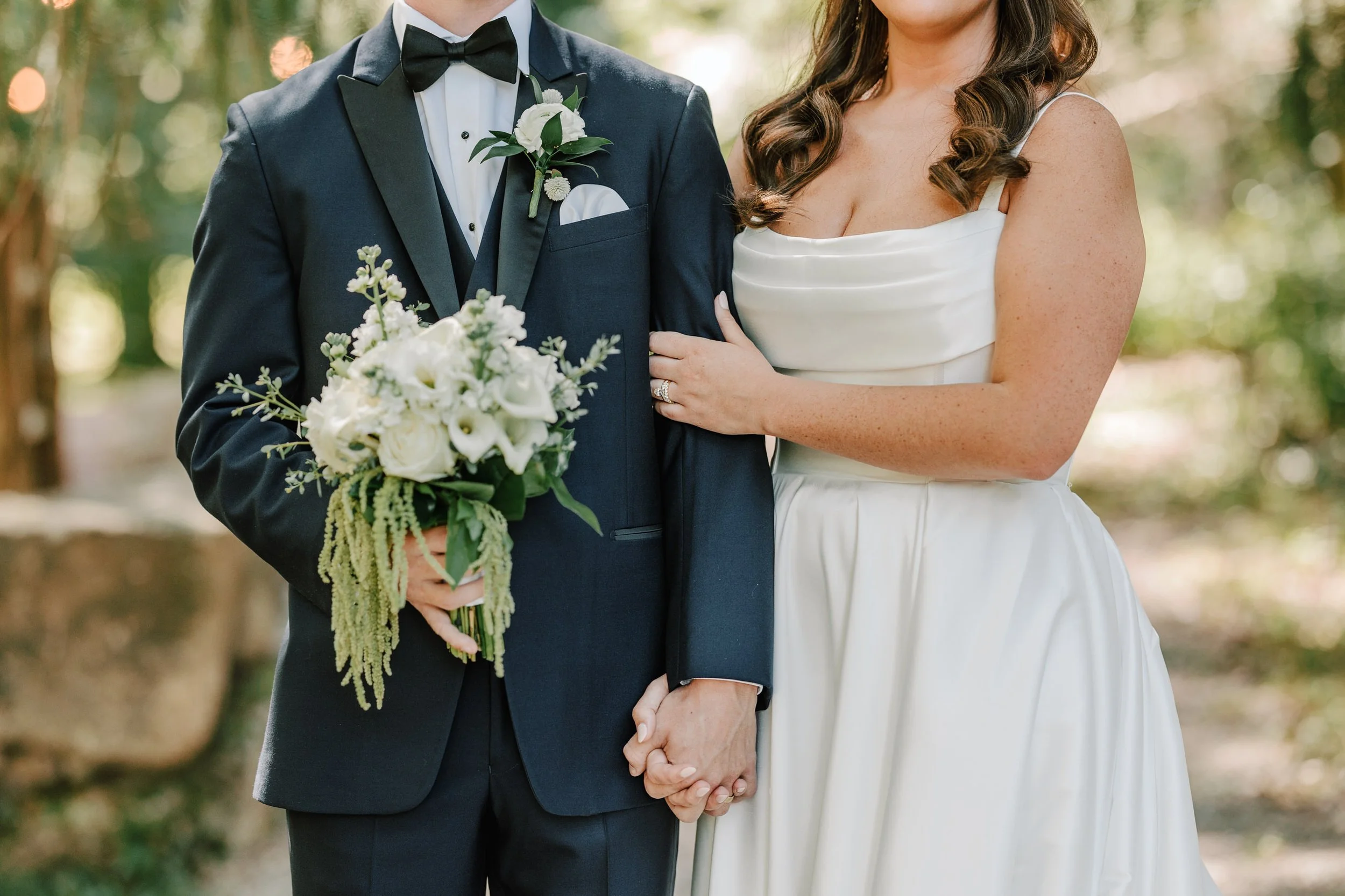 A newly married couple holding hands during their wedding, with the groom in a black tuxedo and the bride in a white satin dress, holding a bouquet of white flowers.