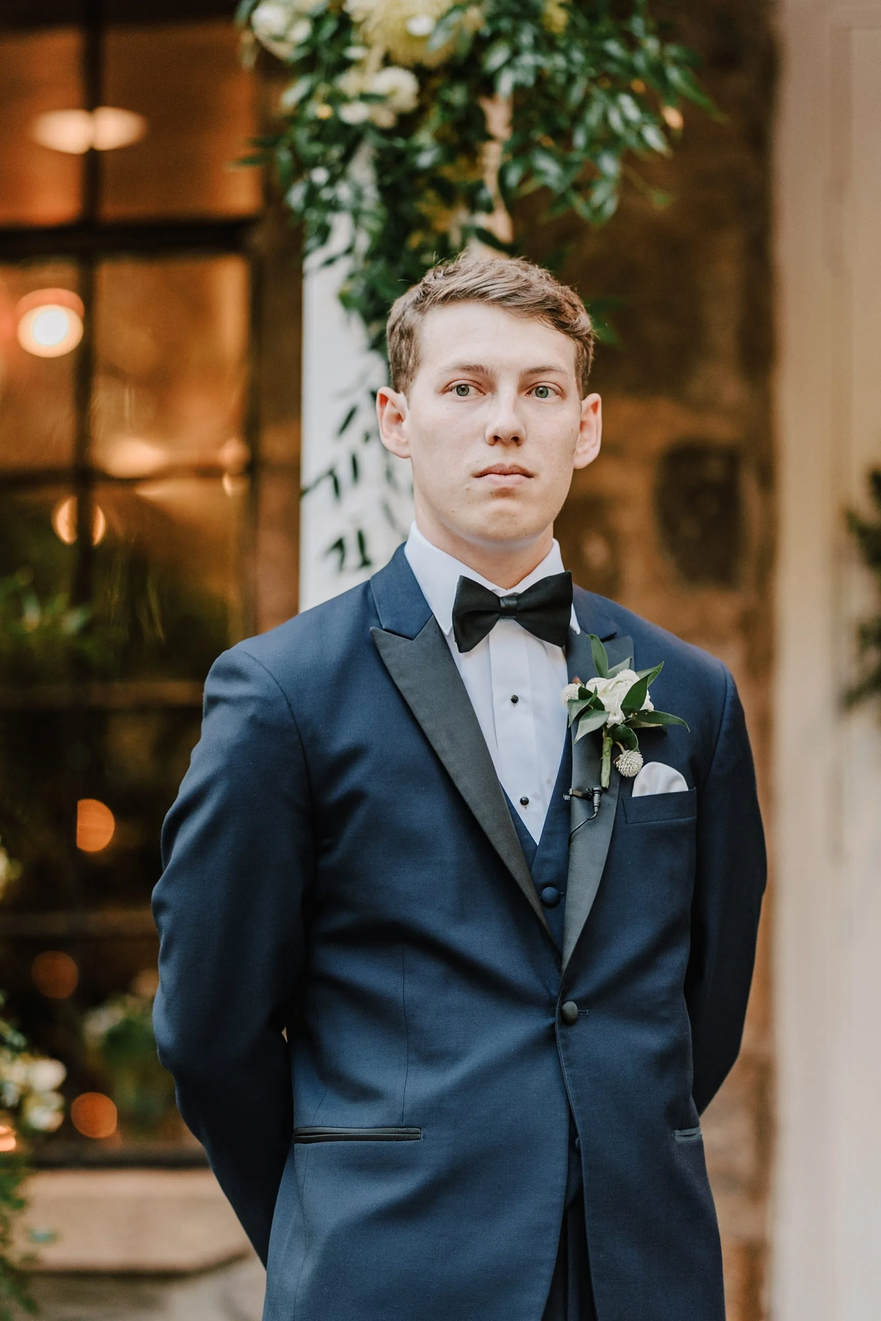 A young man in a navy tuxedo with a black bow tie and boutonniere stands indoors, likely at a formal event or wedding, with a decorative floral arrangement behind him.