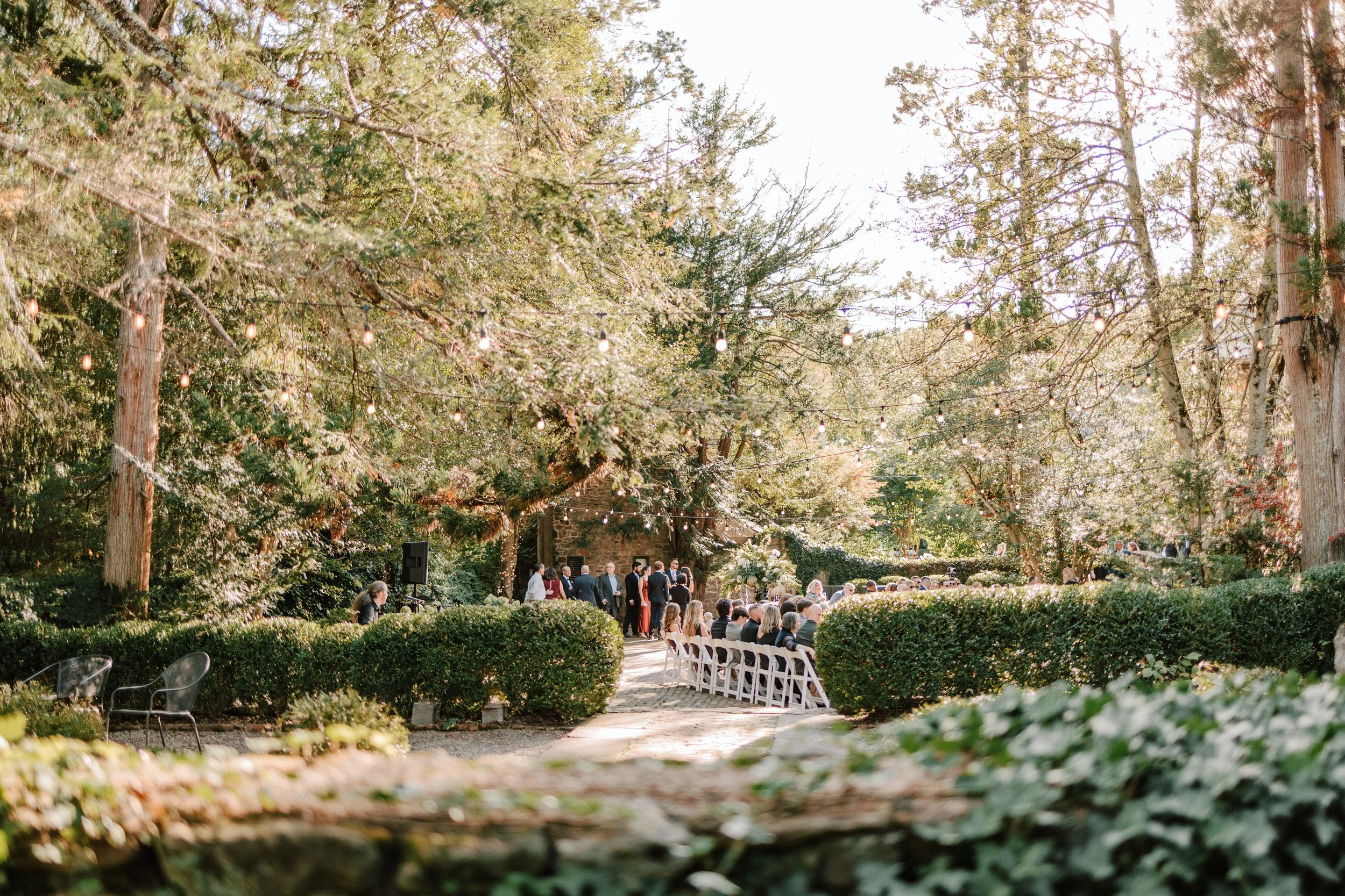 An outdoor wedding ceremony with guests seated on white chairs amidst lush greenery, under string lights and surrounded by tall trees.