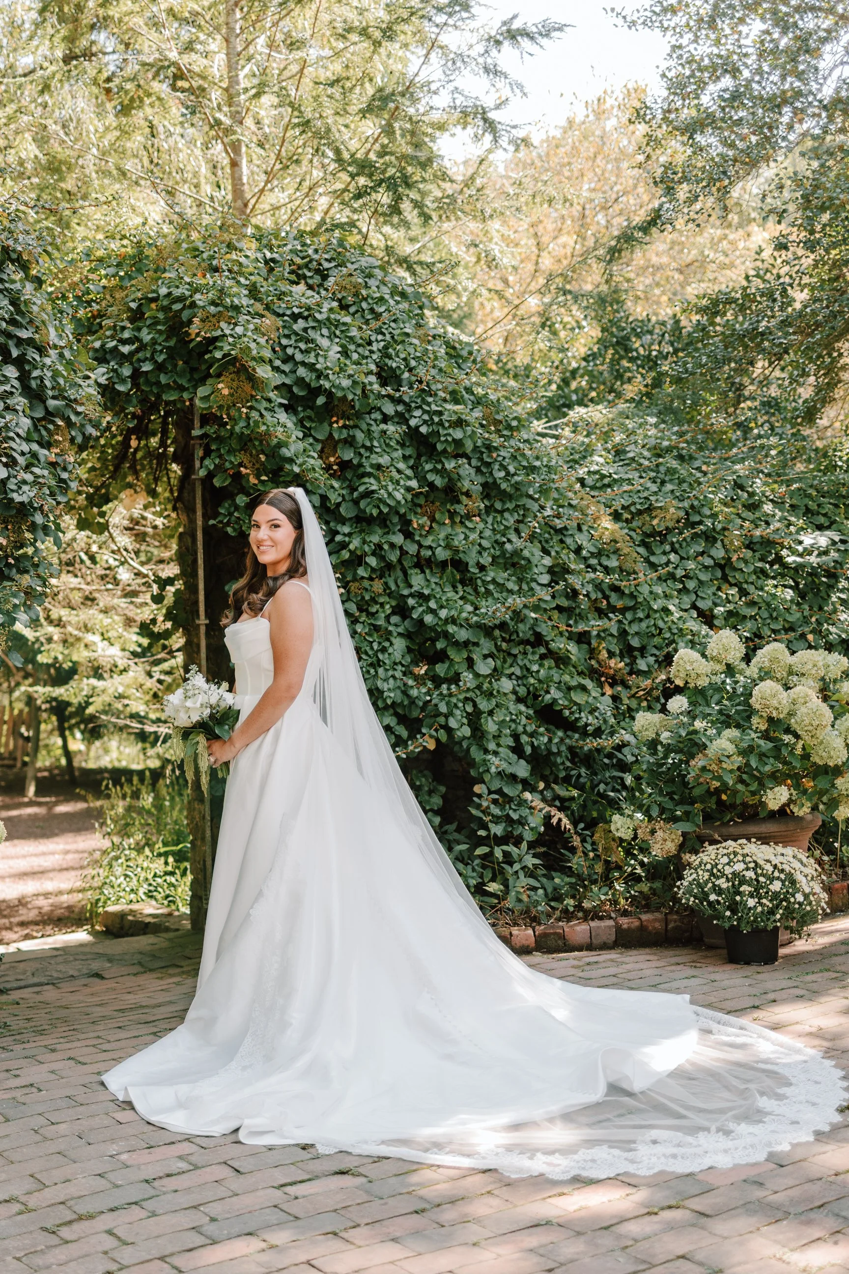 A bride in a white wedding dress with a long train and veil standing outdoors on a brick path, holding a bouquet, surrounded by greenery and white flowers.