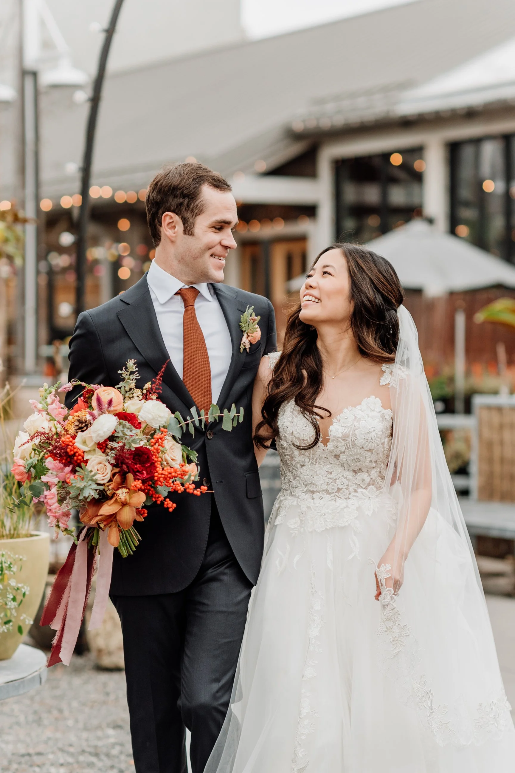 A newly married couple stands outdoors, smiling at each other. The groom wears a dark suit with a white shirt and brown tie, holding a colorful bouquet of flowers. The bride is in a white lace wedding dress with a veil, looking happily at the groom.