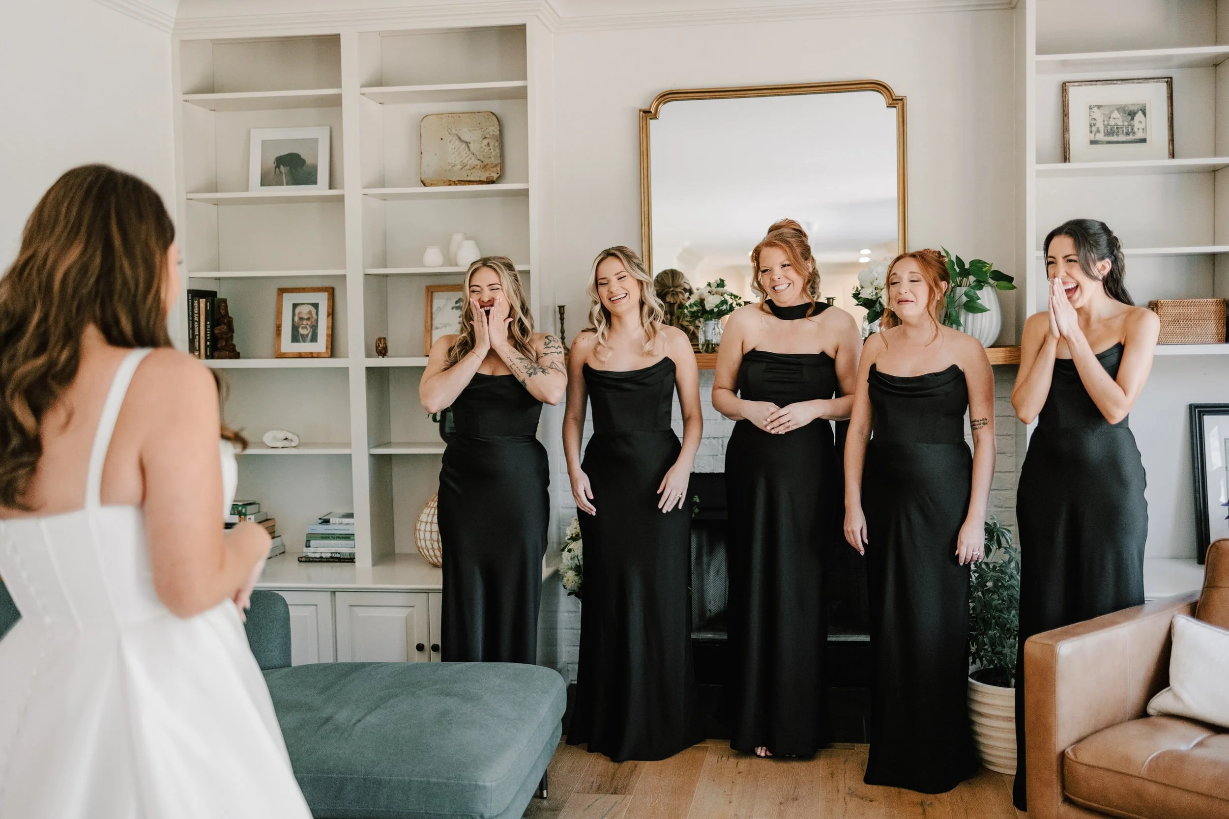 A bride with long brown hair in a white wedding dress is looking at five bridesmaids in black dresses, who are standing in front of a white fireplace with a large mirror and decorative items, all smiling and appearing emotional.