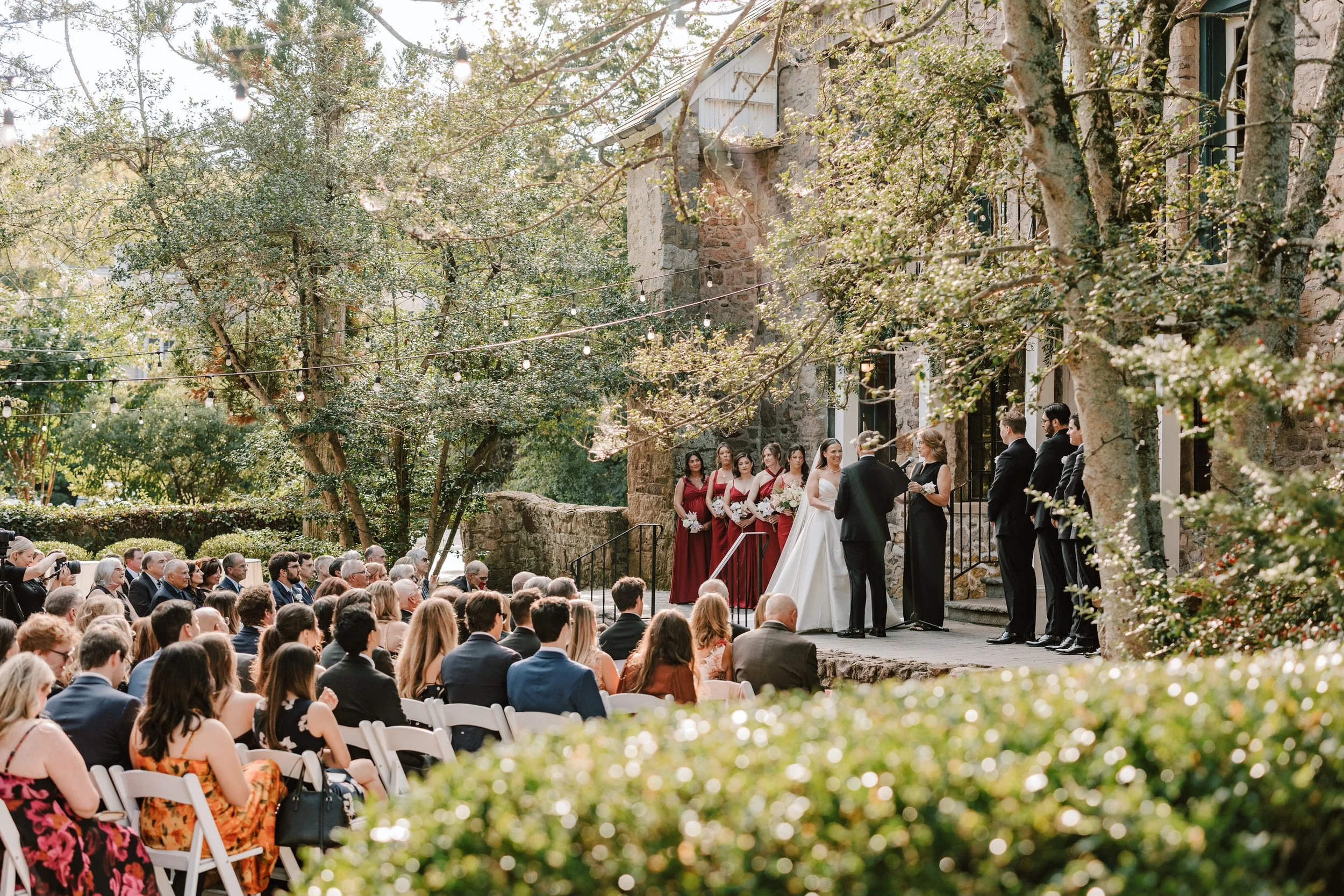 Outdoor wedding ceremony with bride and groom at altar, surrounded by bridesmaids in burgundy dresses and groomsmen in black suits, guests seated in chairs, trees, and string lights in the background.