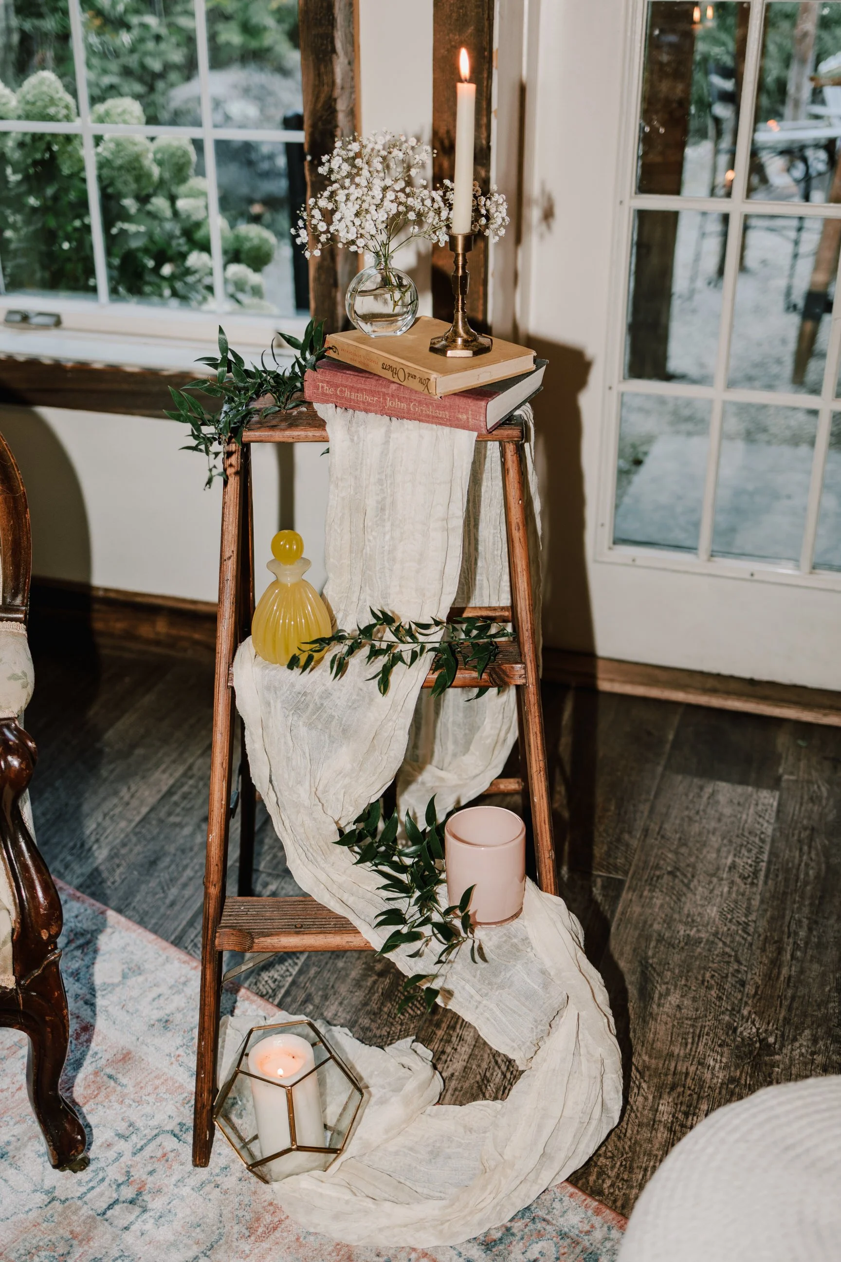 Decorative centerpiece on a wooden step ladder with candles, books, vases, and greenery, near a window and door.