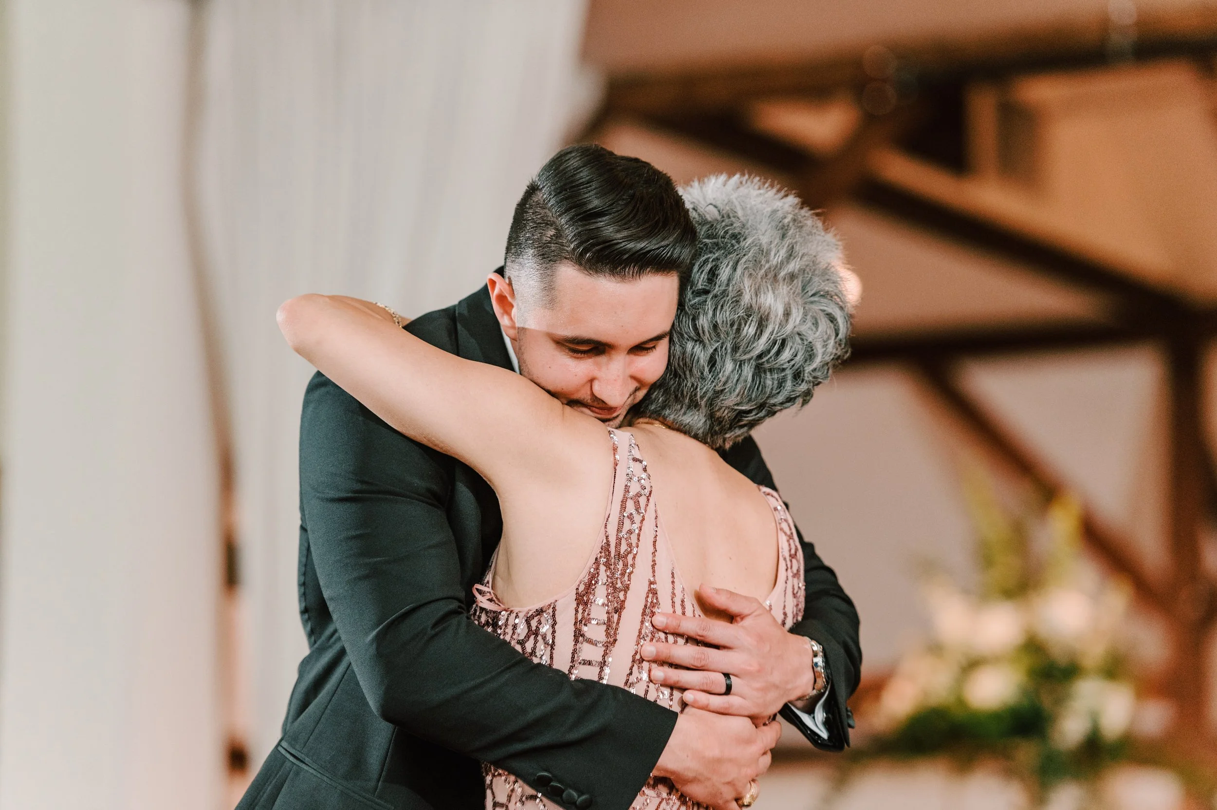 A young man in a black suit and a woman with gray hair and a pink dress are hugging during a celebration or event.