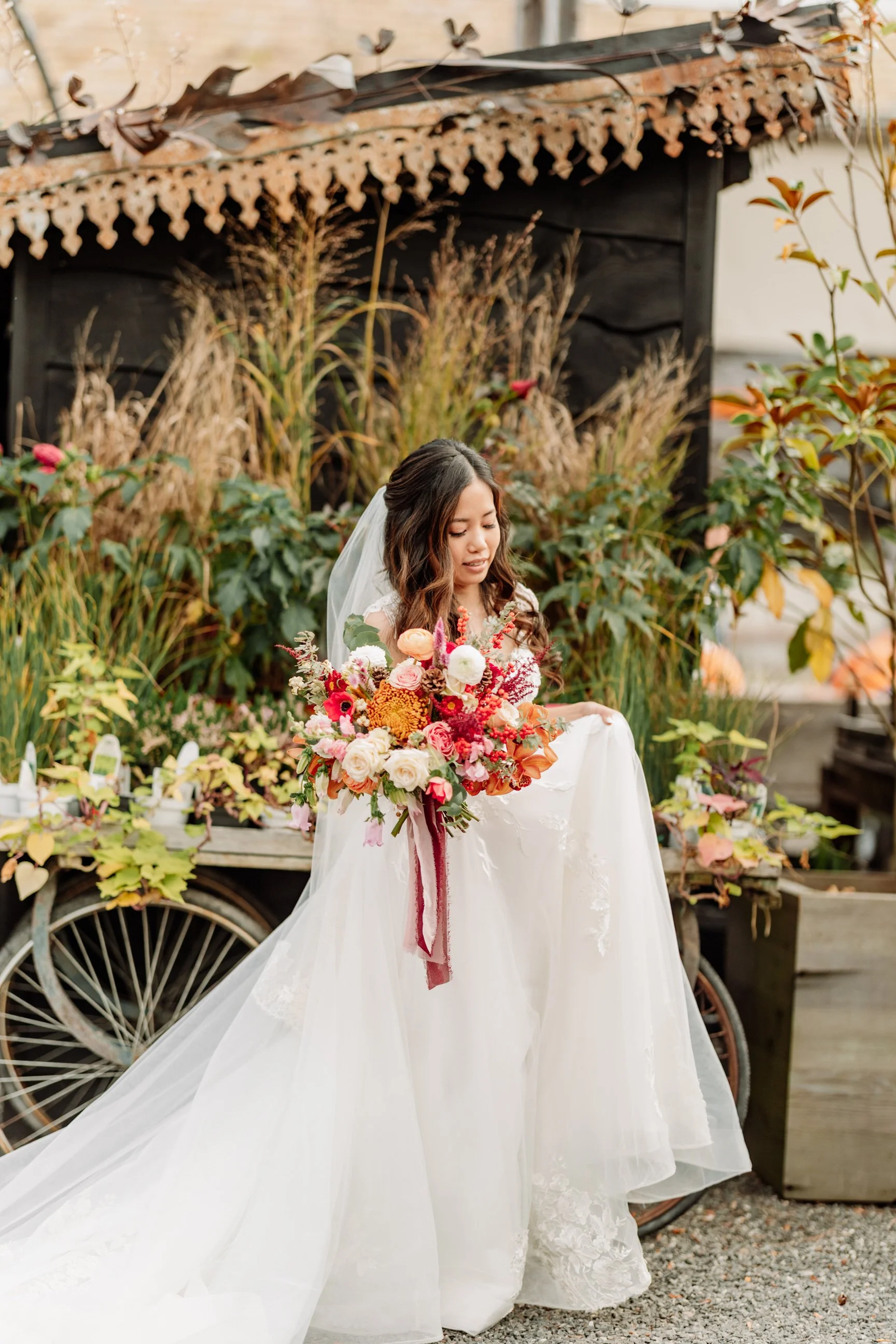 Bridal portrait of a woman in a white wedding gown holding a large colorful bouquet of flowers, standing outdoors in front of a rustic wooden cart with plants and a black wooden shed.