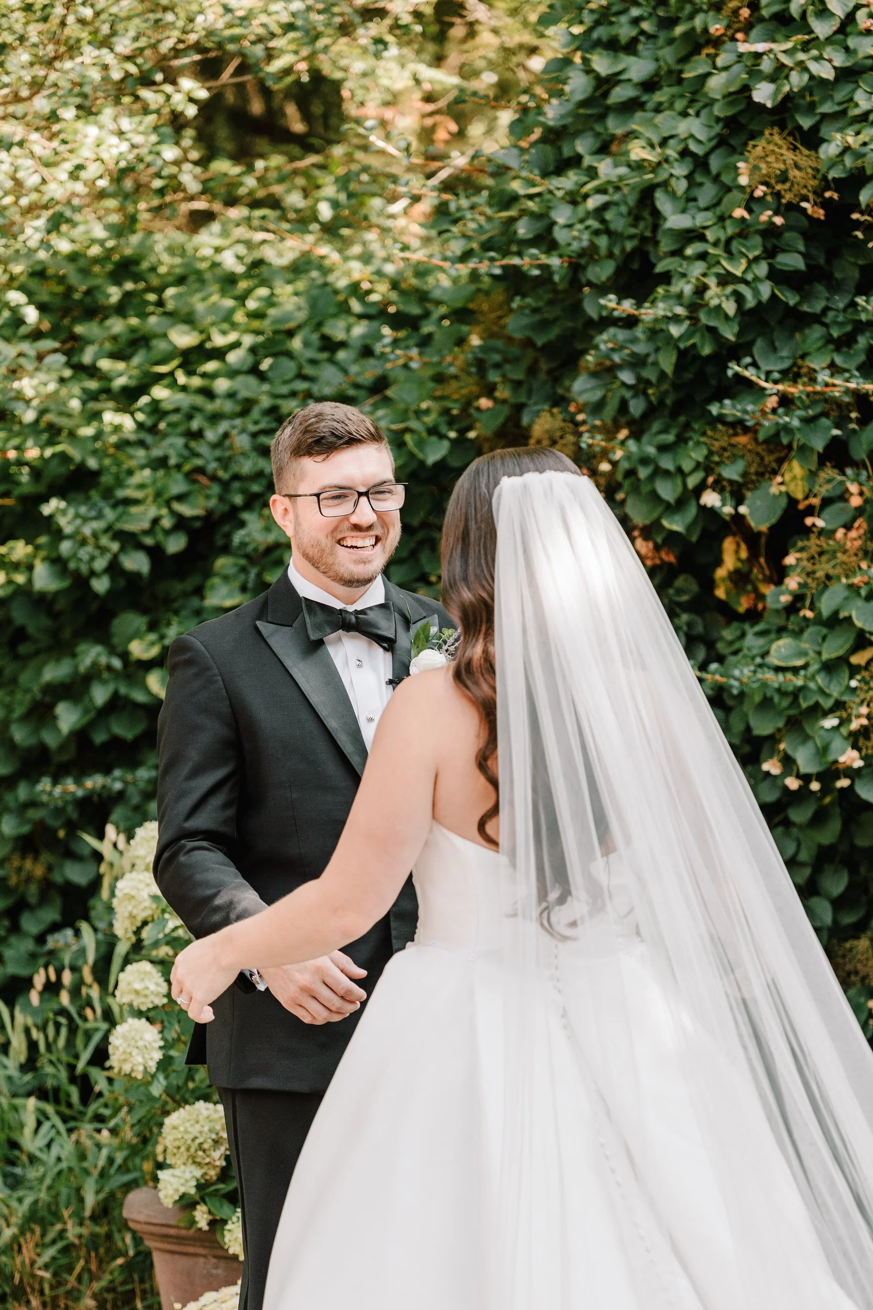 A bride and groom exchange vows outdoors during their wedding ceremony surrounded by lush green foliage.