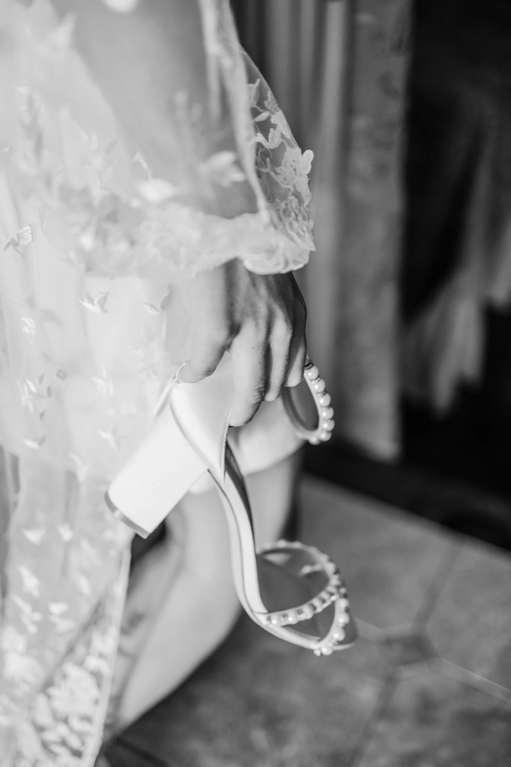 A hand holding a pair of pearl-embellished sunglasses. The hand is partially covered by lace and fabric, suggesting the person is wearing a wedding dress or formal attire.