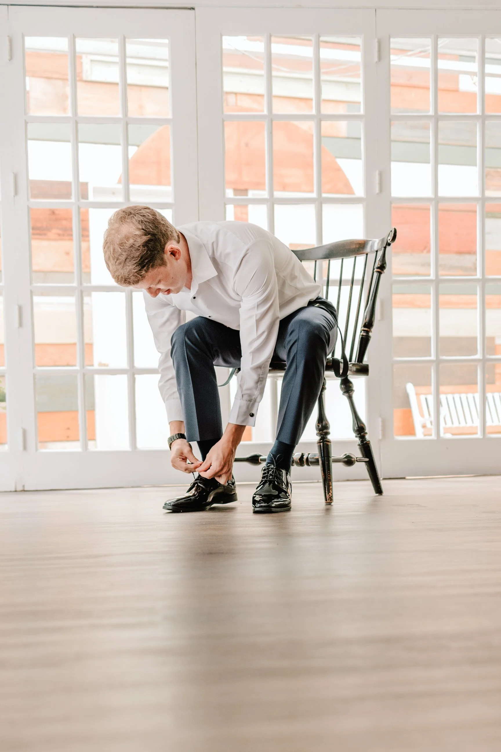 Young man in white shirt and dark pants sitting on a wooden chair, tying his black dress shoes near large windows with multiple glass panes.