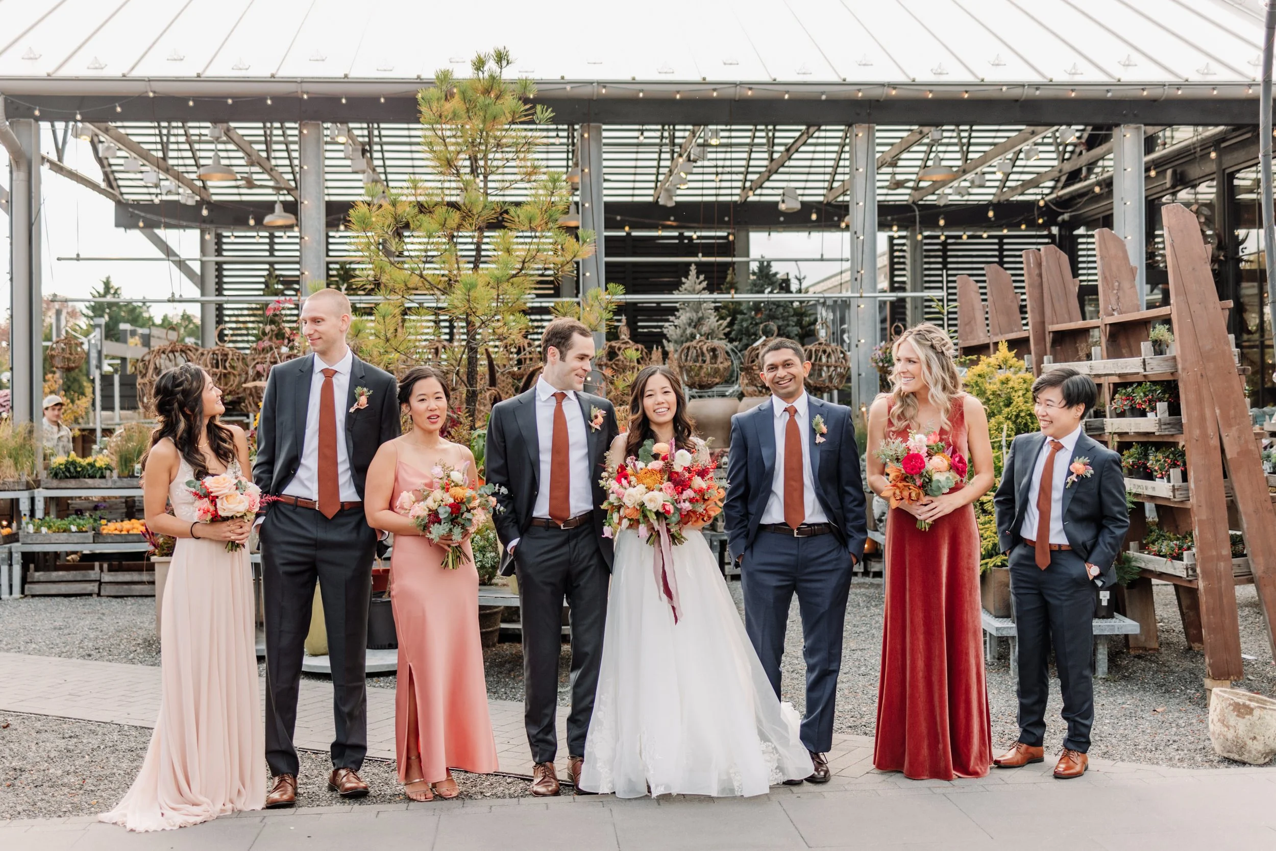 Group of eight people, including a bride in a white wedding dress and seven wedding guests, standing together outdoors at a garden center, holding bouquets of flowers, with smiling and talking to each other.