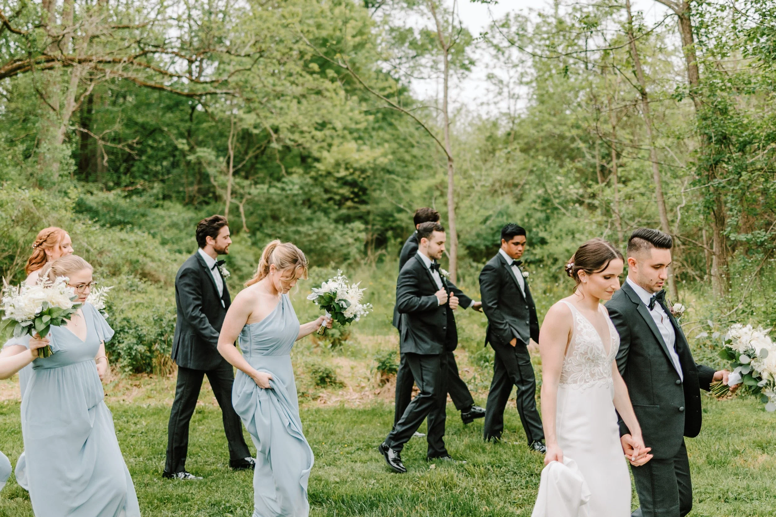 A wedding procession walking outdoors in a wooded area, with bridesmaids in light blue dresses holding bouquets and groomsmen in black tuxedos walking behind the bride and groom in a white dress and black tuxedo.