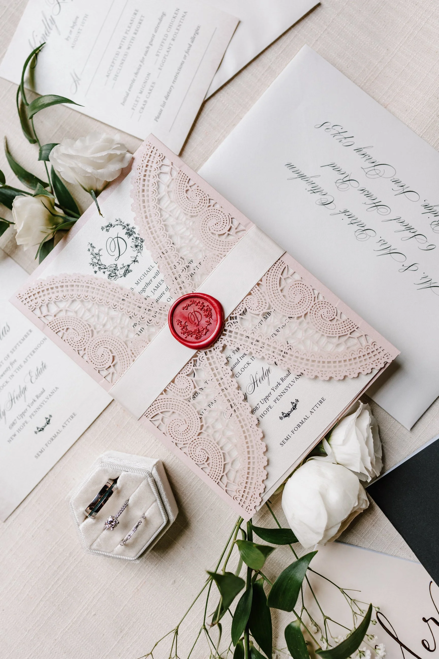 Wedding invitations with lace details, white flowers, and a red wax seal, along with engagement or wedding rings in a white ring box on a light-colored table.