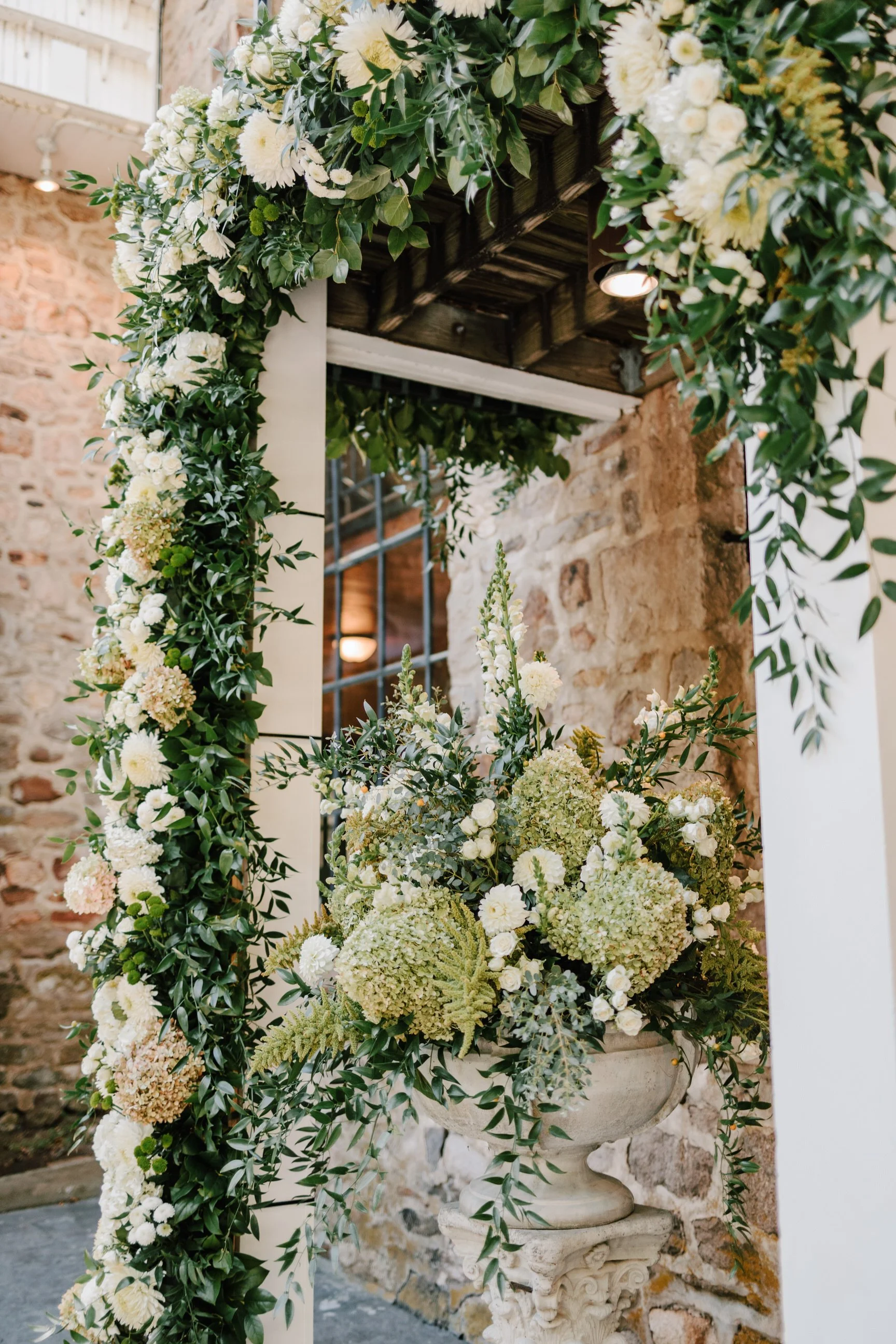 Decorative floral arrangement with white and pale green flowers and green foliage in a stone urn, set against a rustic brick wall with an archway.