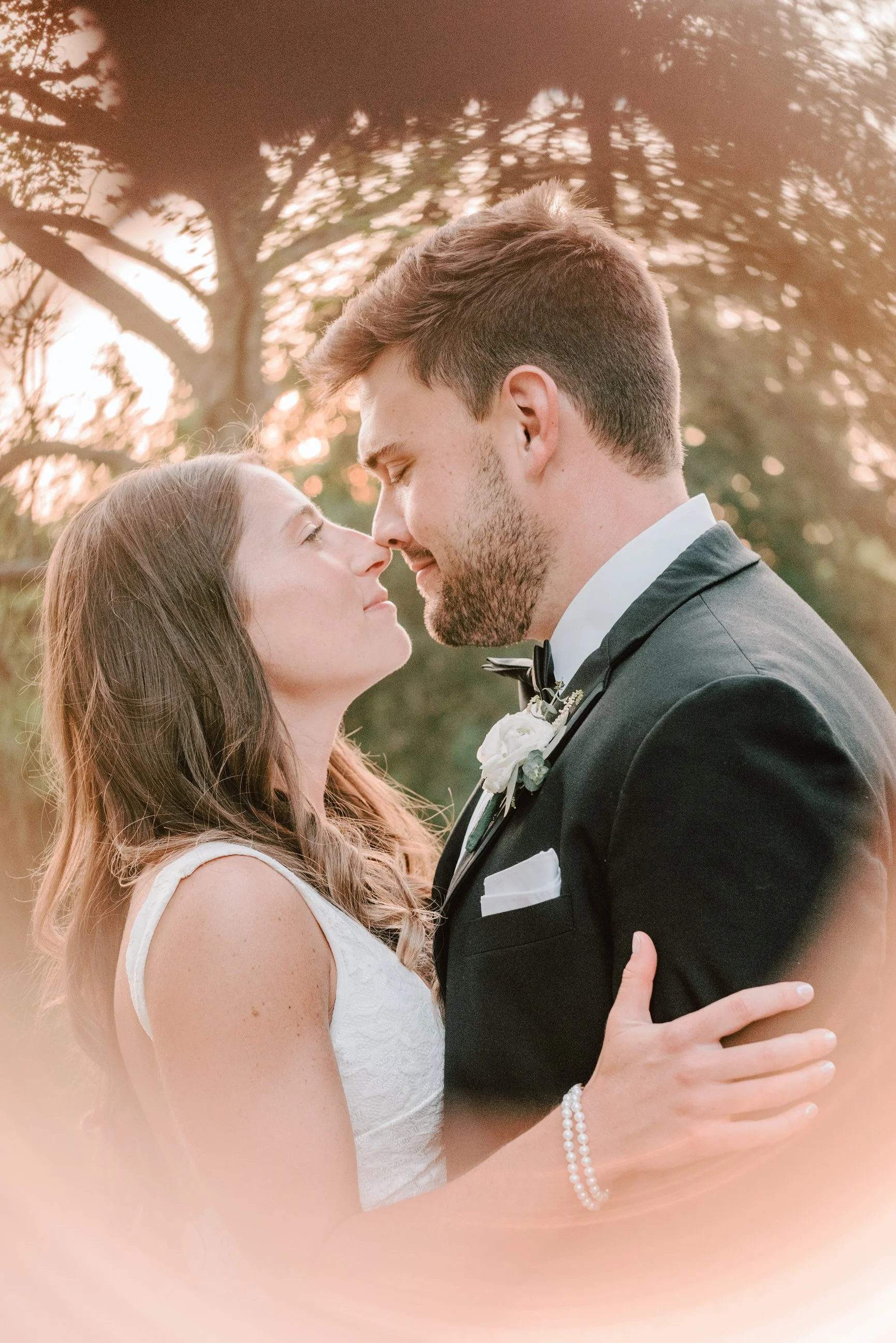 A bride and groom close together outdoors, about to kiss, with warm sunset lighting and trees in the background.