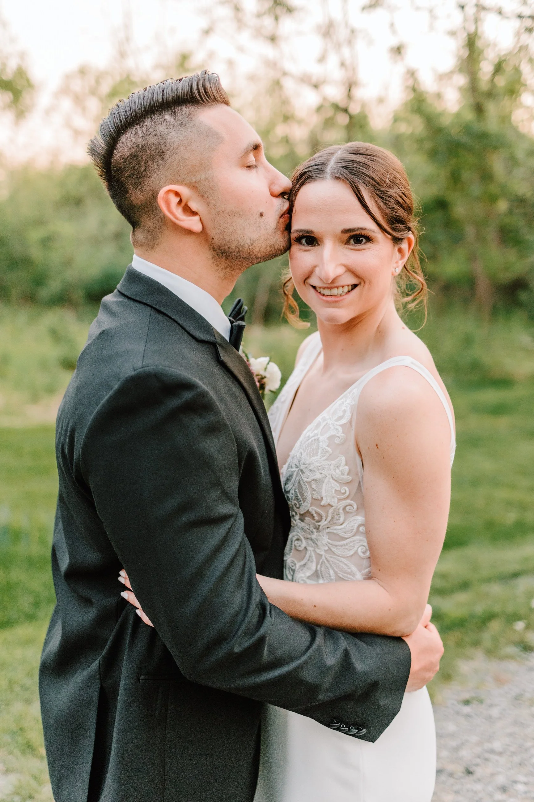 A groom in a black tuxedo and a bride in a white wedding dress embracing outdoors, with the groom kissing the bride on her forehead and the bride smiling at the camera.