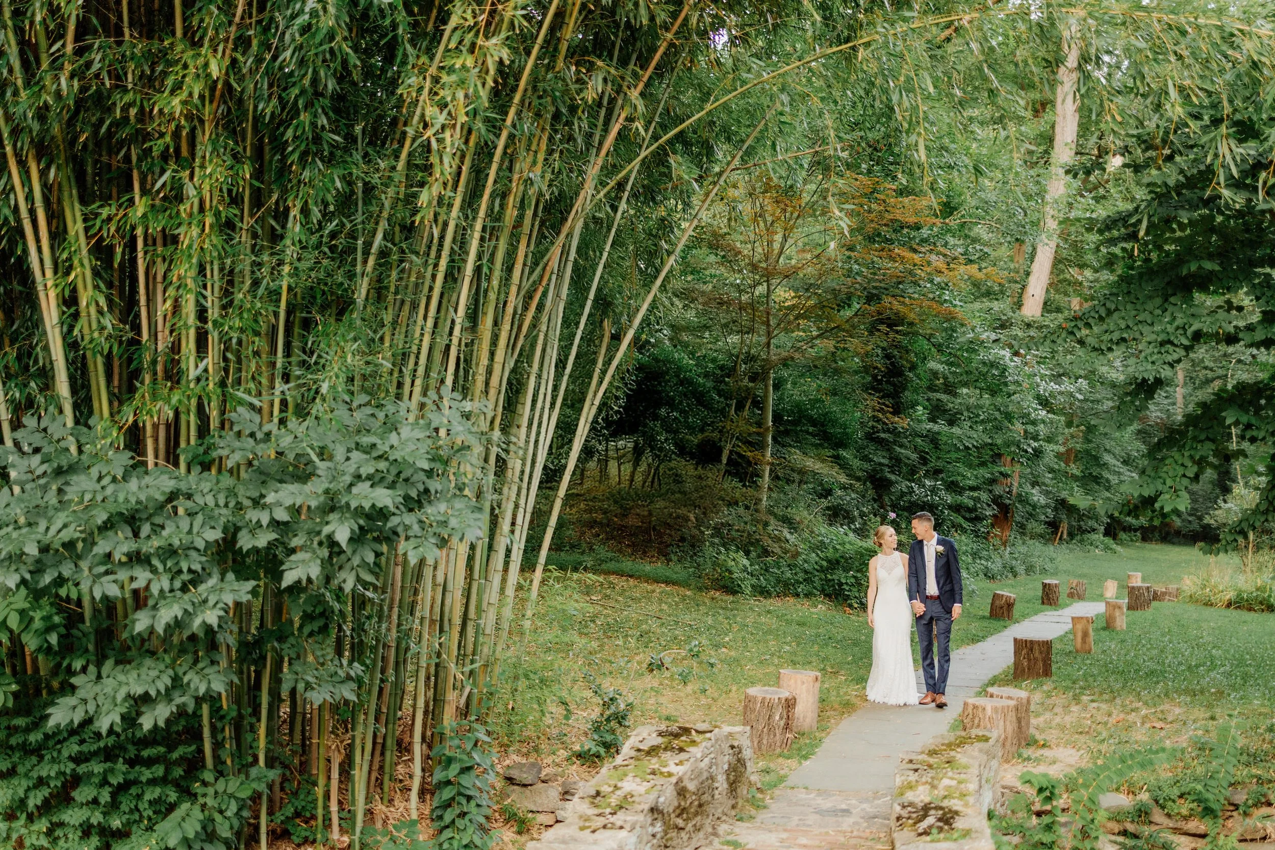 Couple in wedding attire walking on a path surrounded by lush greenery and bamboo.