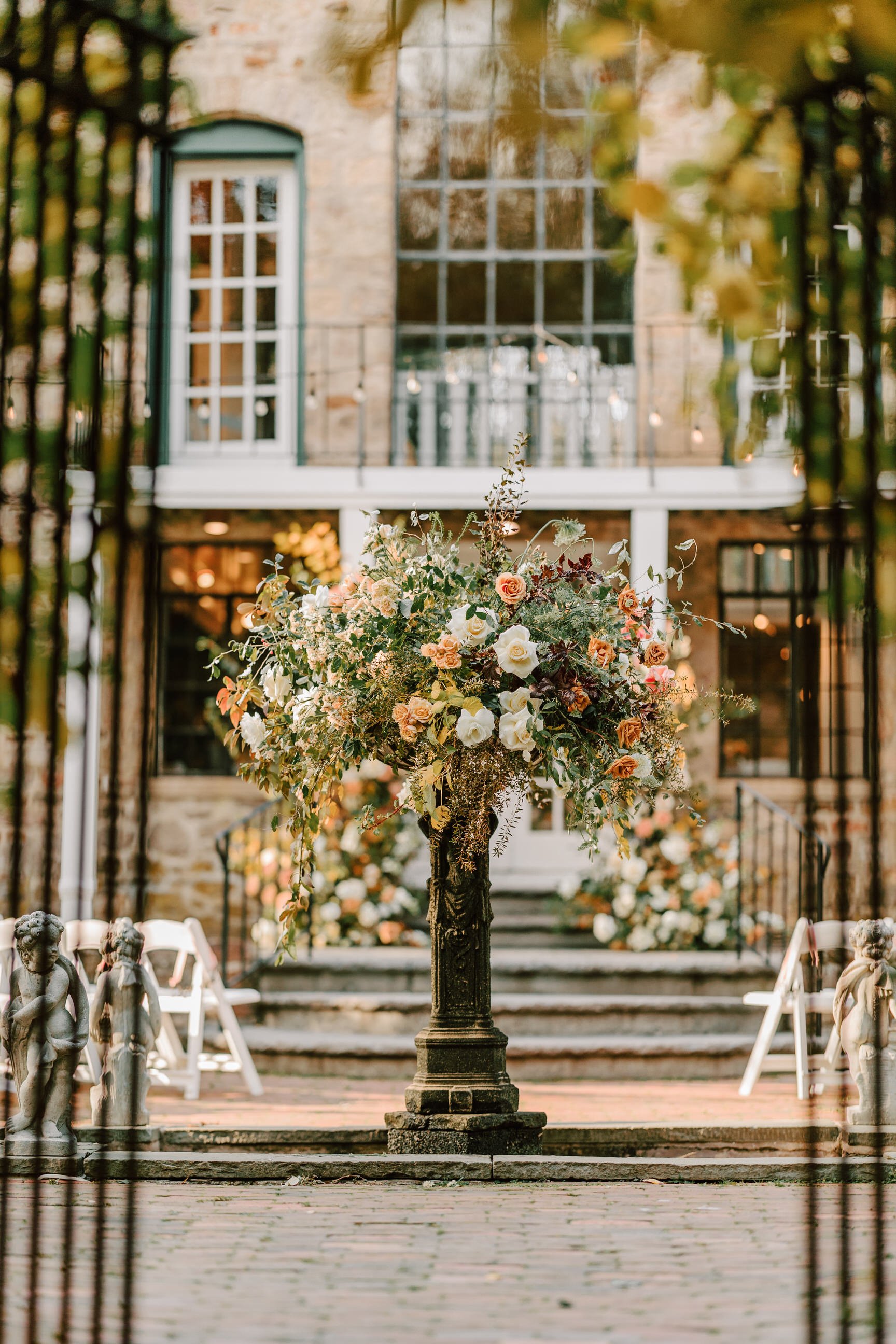 A large floral arrangement with white and peach roses on a pedestal, set outdoors with steps and a stone building with large windows in the background, decorated for a wedding or special event.