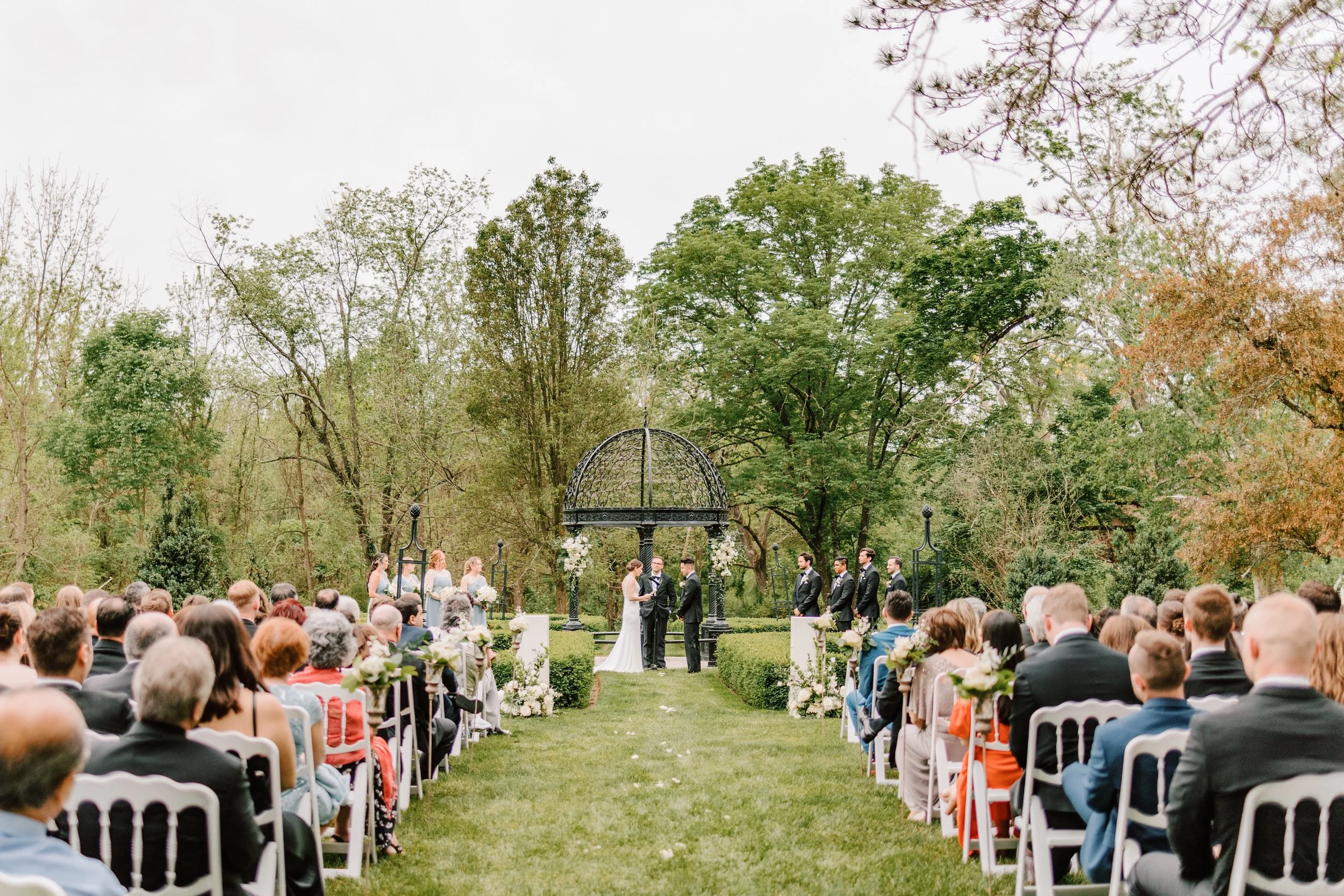 A wedding ceremony outdoors with a bride and groom exchanging vows under a black gazebo, surrounded by guests and lush trees.