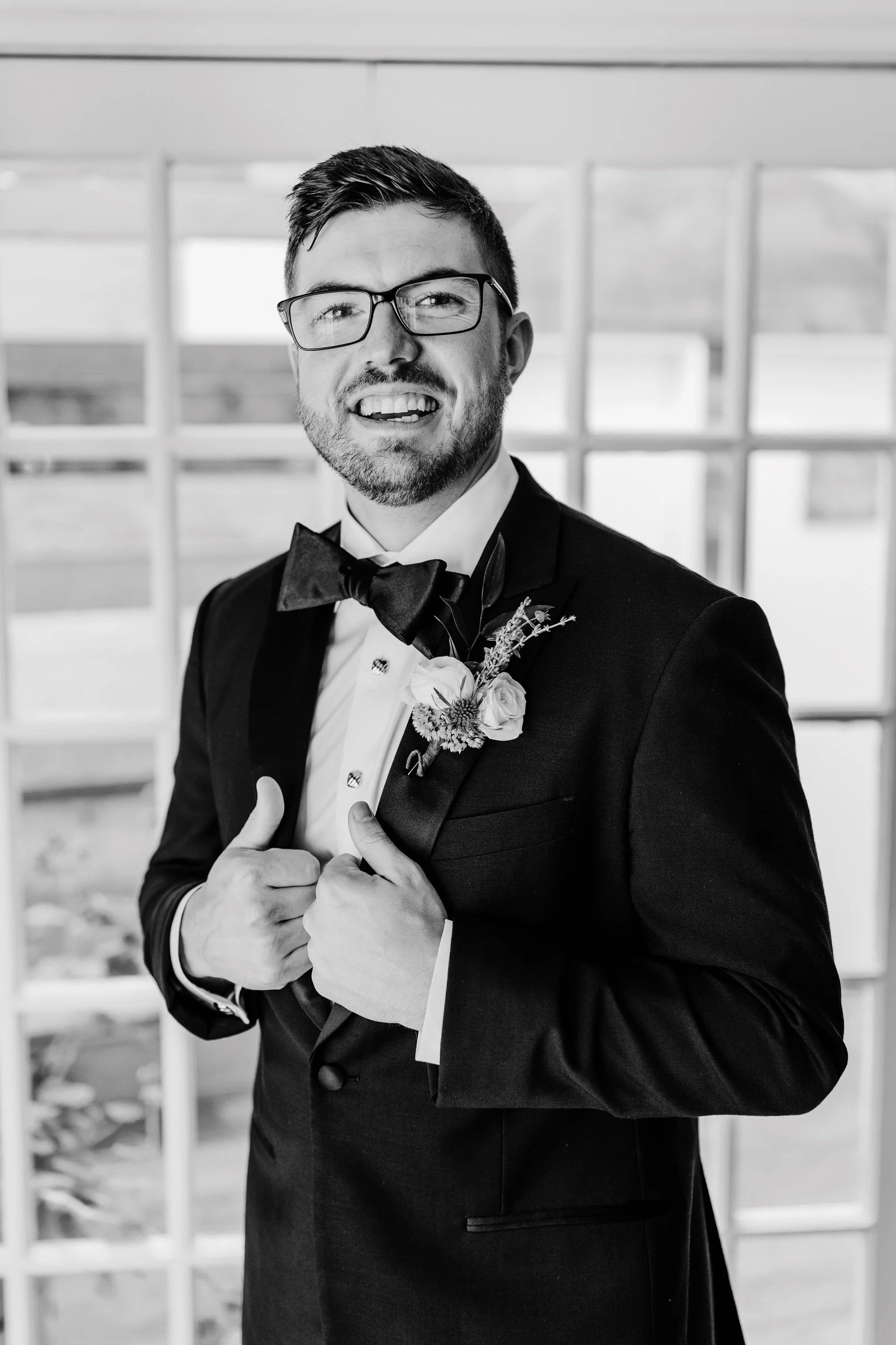 A man in a tuxedo with a bow tie and glasses, smiling, adjusting his tuxedo jacket, in front of a window.