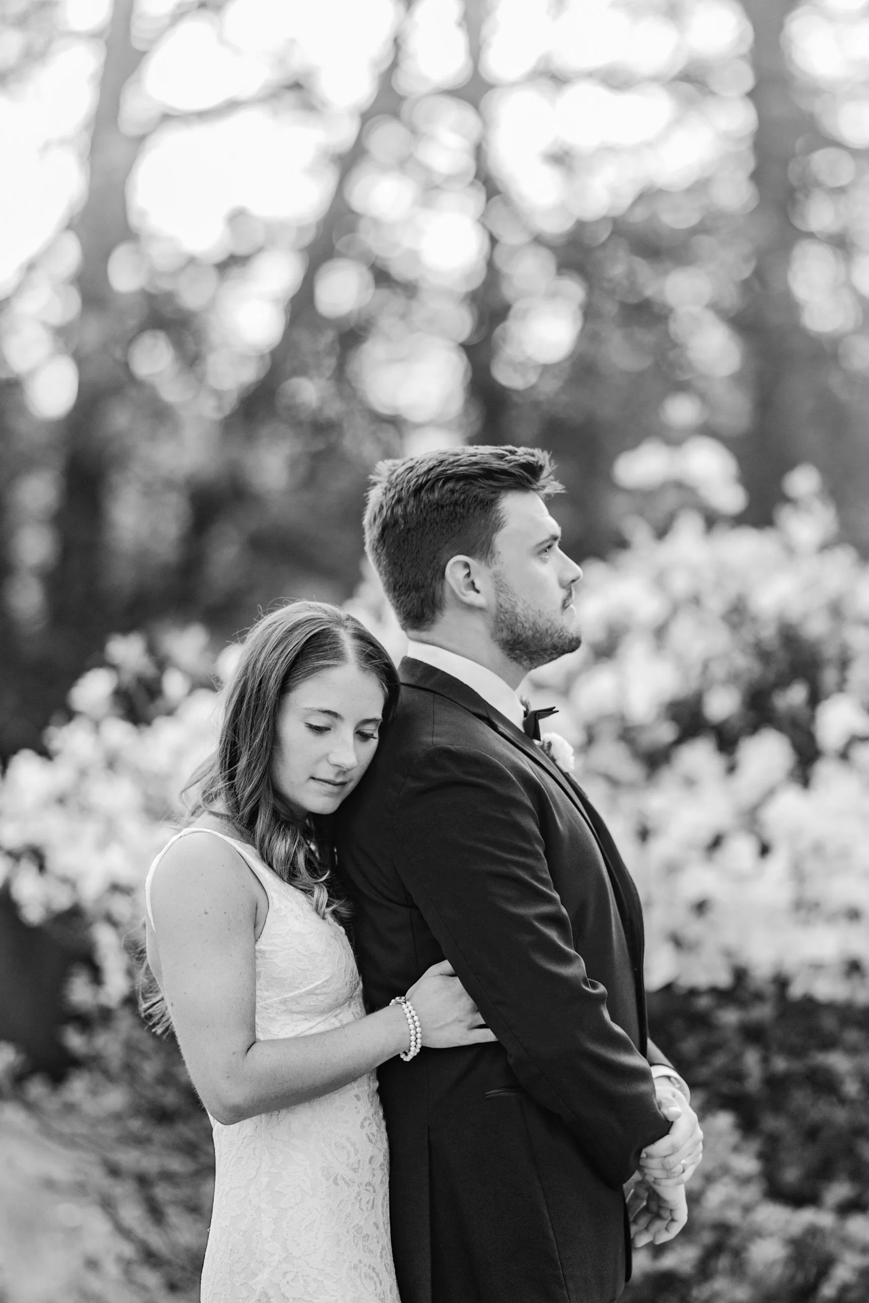 A black and white photo of a bride and groom standing outdoors, with the bride resting her head on the groom's shoulder. The groom is dressed in a tuxedo with a bow tie, and the bride is wearing a lace wedding dress. They are surrounded by blurred tr