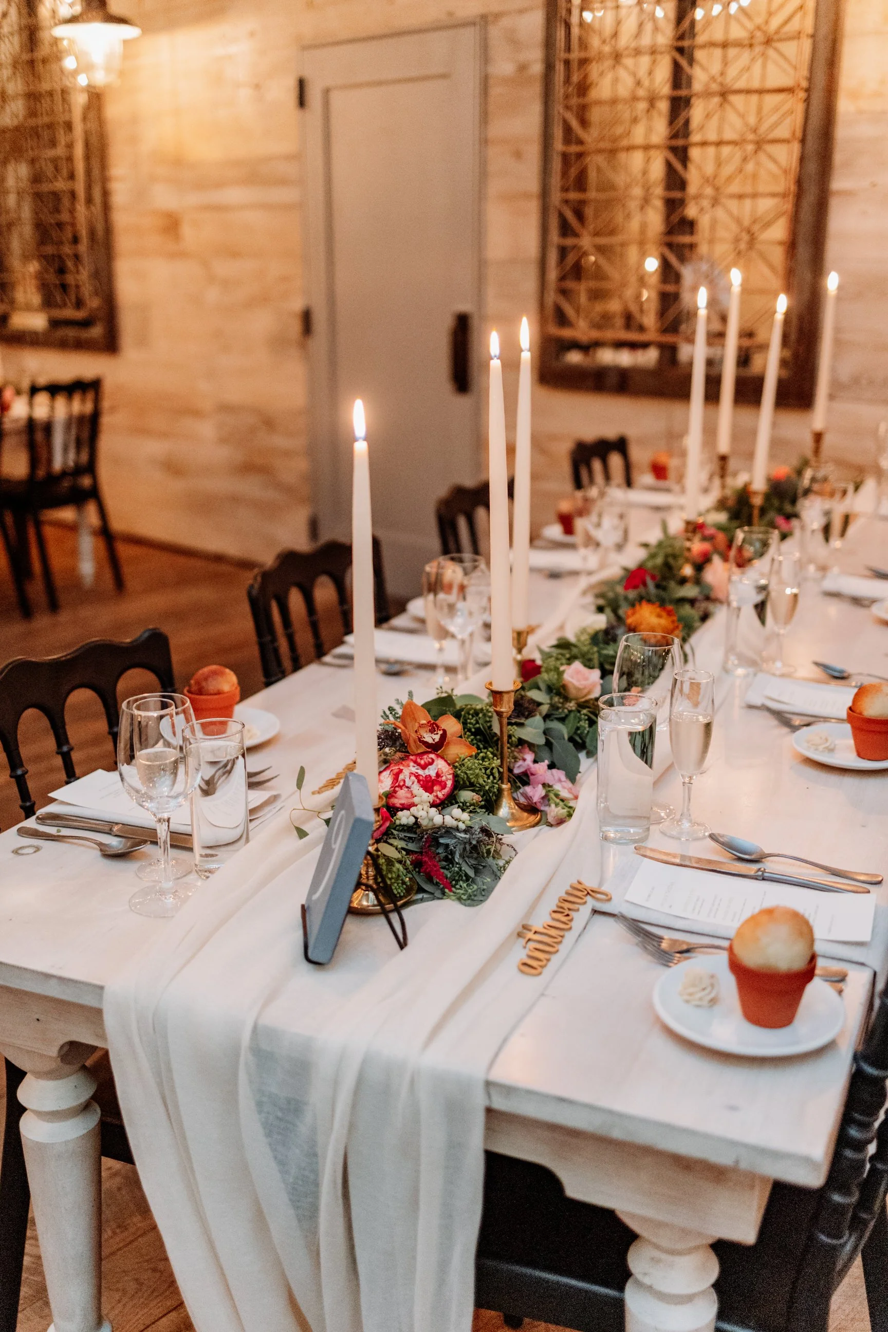 A decorated wedding or event table with tall white candles, a floral garland, water glasses, and baked goods on small plates in a rustic wood room.
