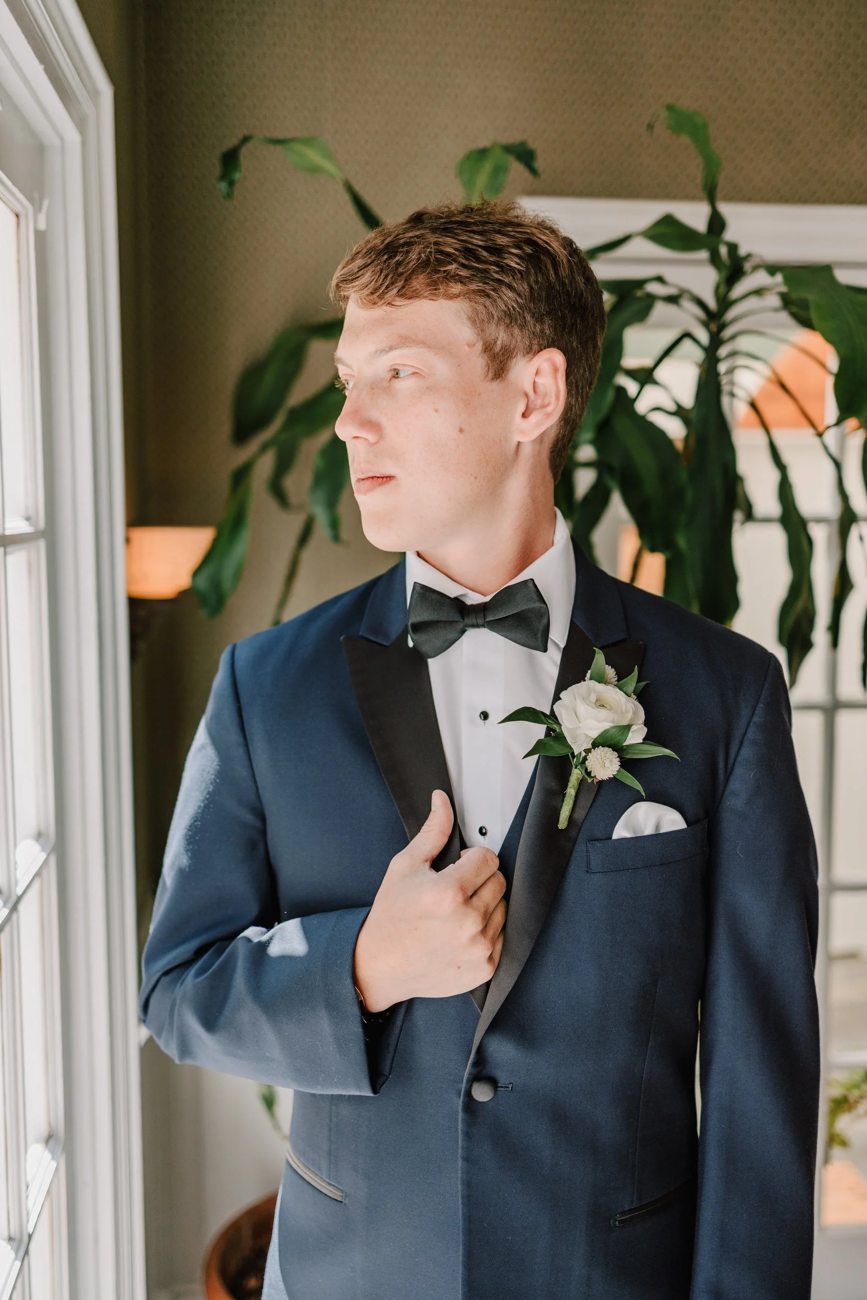 A young man in a navy blue tuxedo with a black bow tie, white shirt, and boutonniere looking out a window.