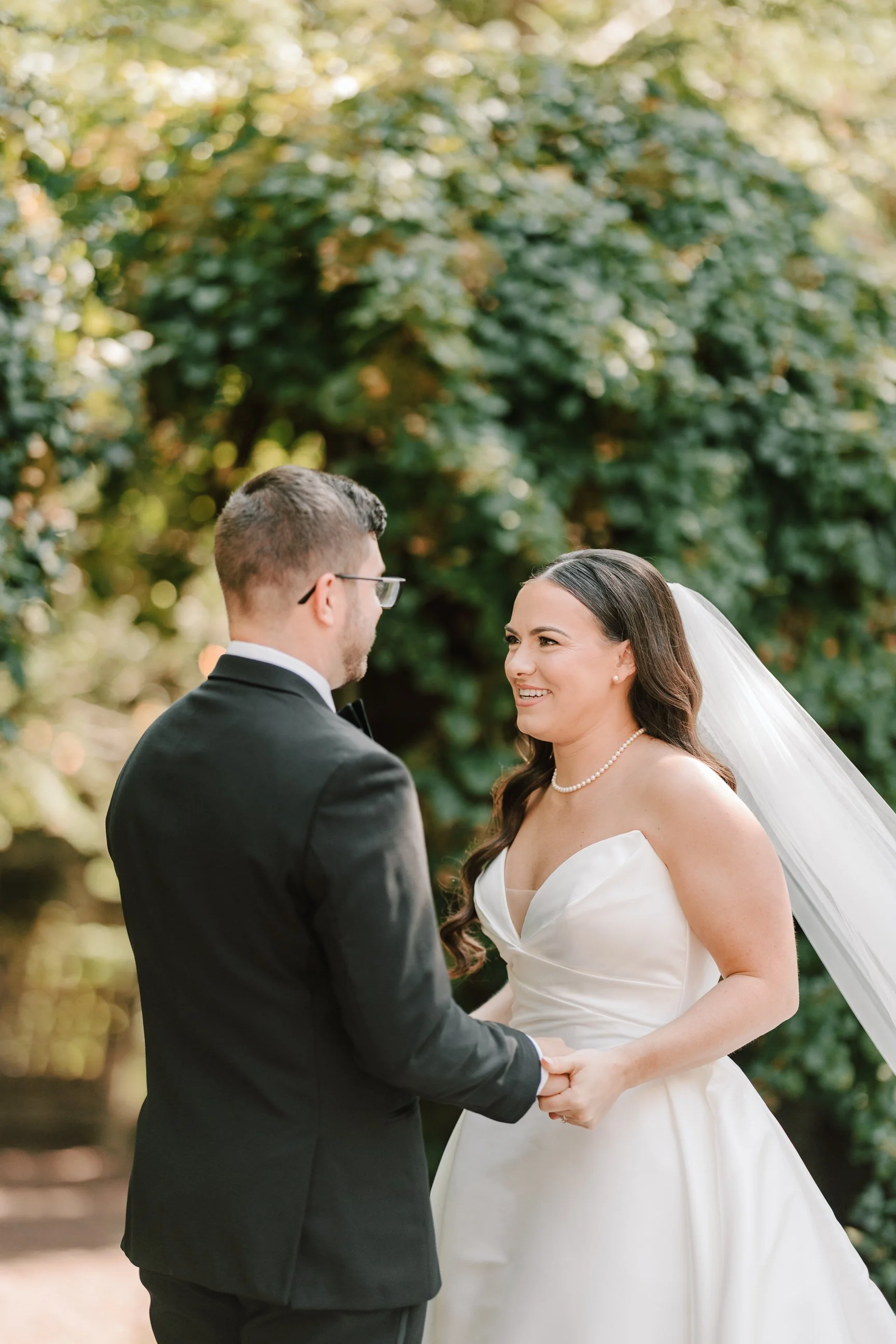 A bride and groom holding hands and smiling at each other outdoors with trees in the background.