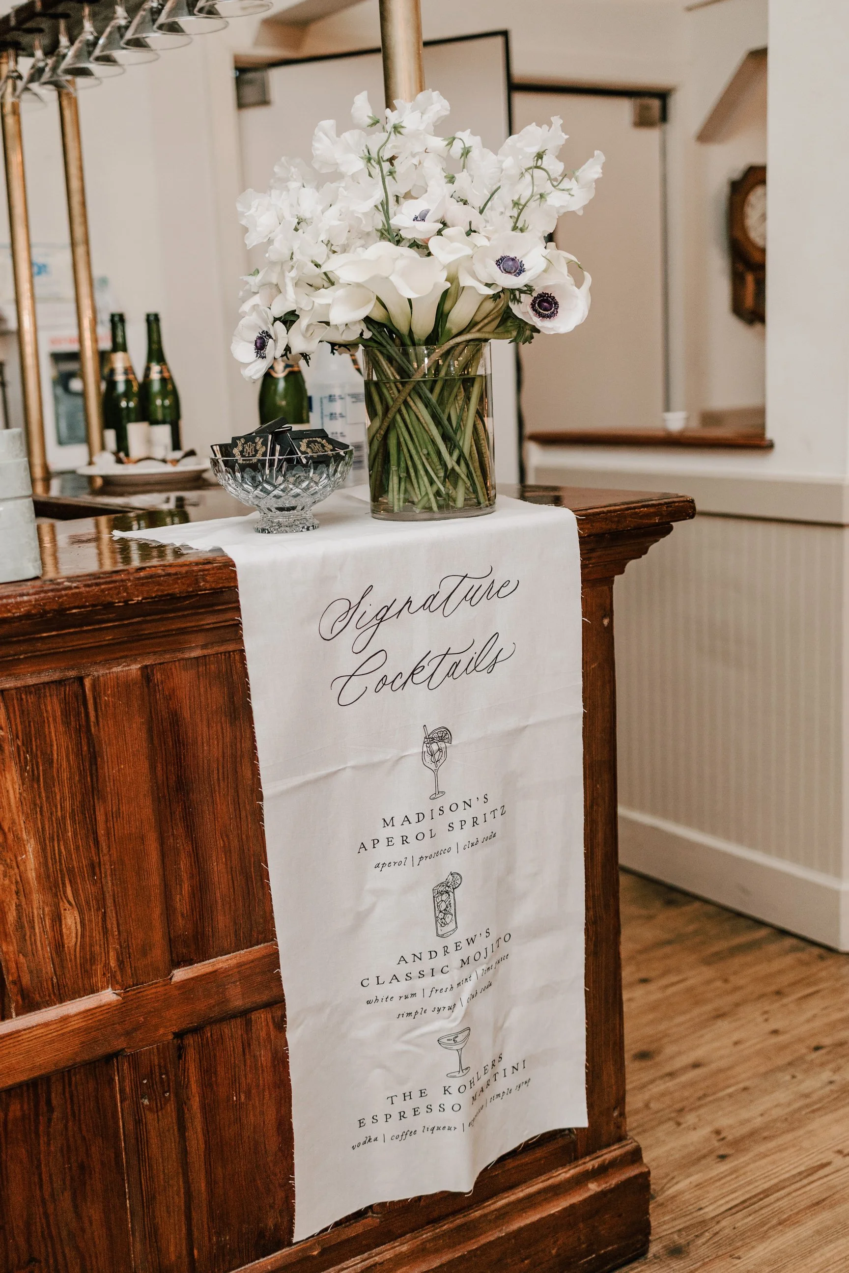 A wooden bar counter with a white cloth sign reading 'Signature Cocktails,' a glass vase with white flowers, and a small glass bowl.