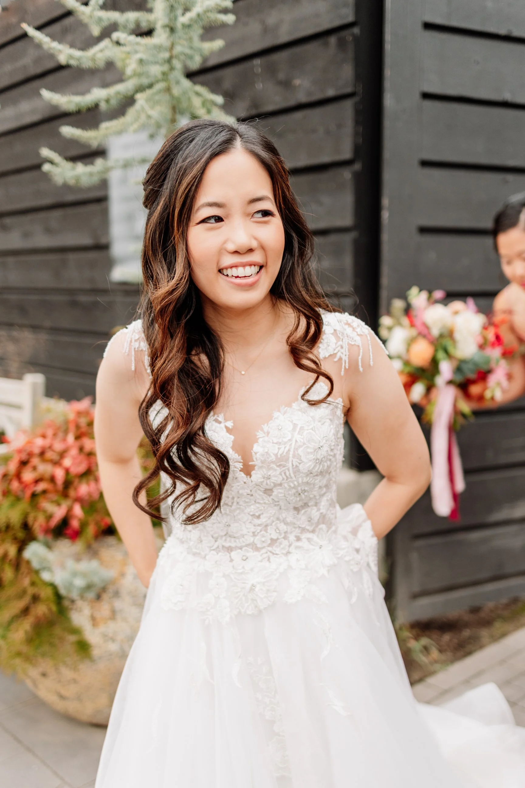 A smiling woman in a wedding dress standing outdoors near a black wooden wall, with flowers and plants in the background.